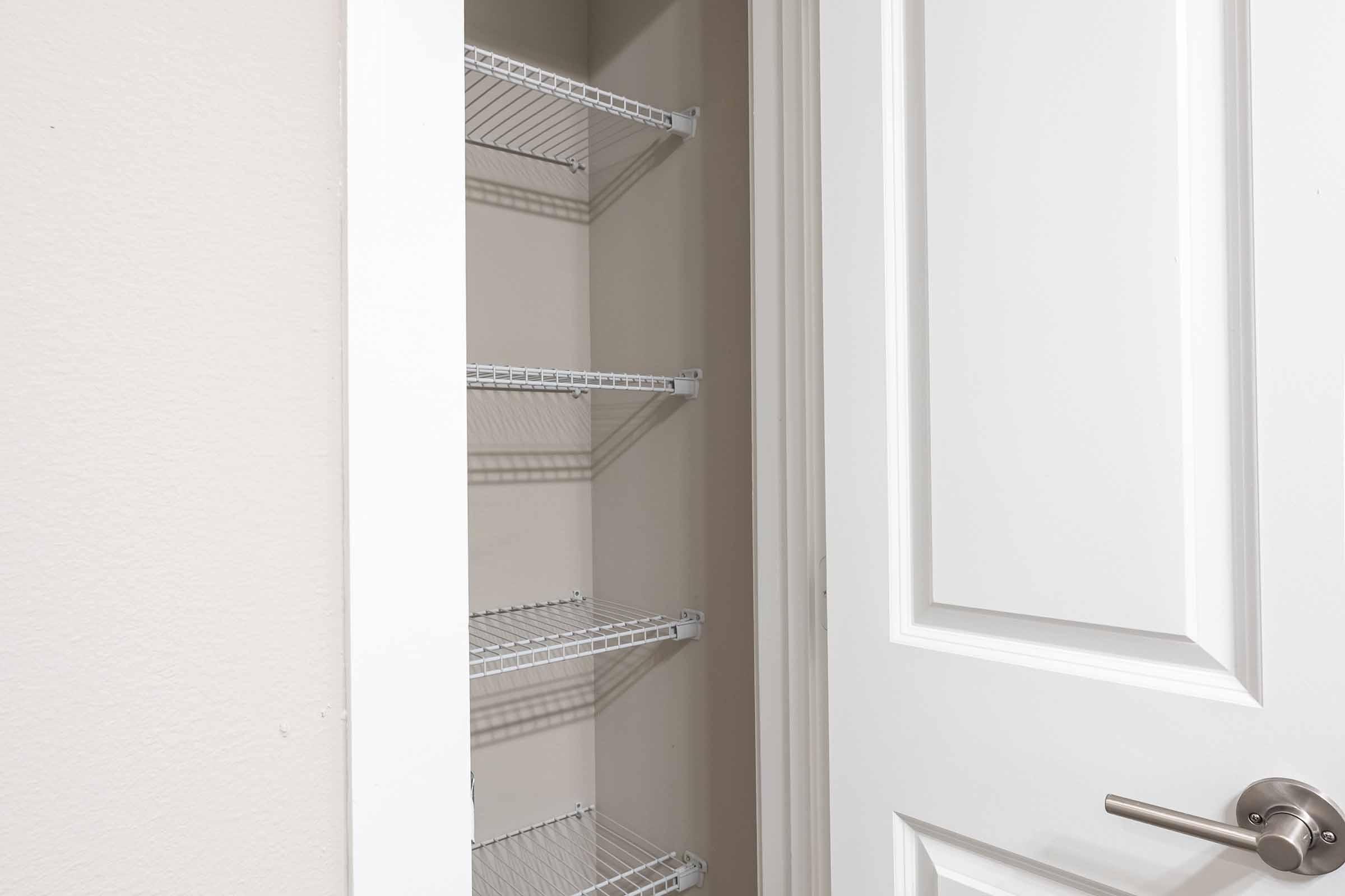 A clean, empty closet with white wire shelves visible. The door is partially open, revealing a plain wall and the shelving inside the storage space. The closet appears to be well-organized and lightly illuminated, with a simple white door and brushed metal handle.