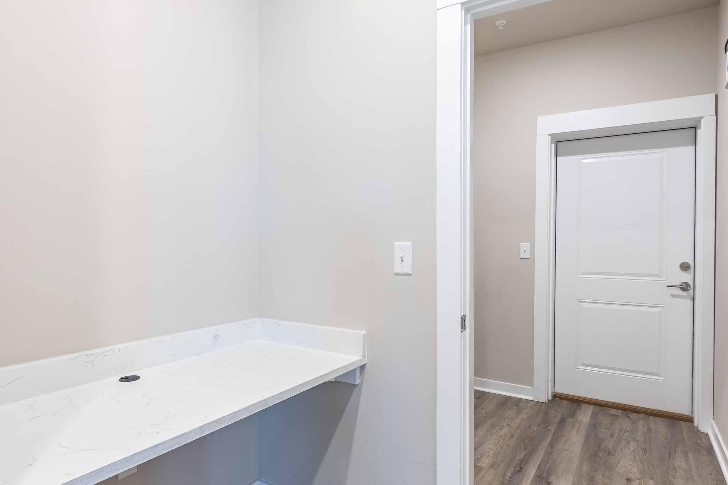 A minimalist interior view of a laundry room, featuring a plain countertop on the left and a white door on the right leading to another space. The walls are light-colored, and the flooring is a light wood finish, creating a bright and open atmosphere.