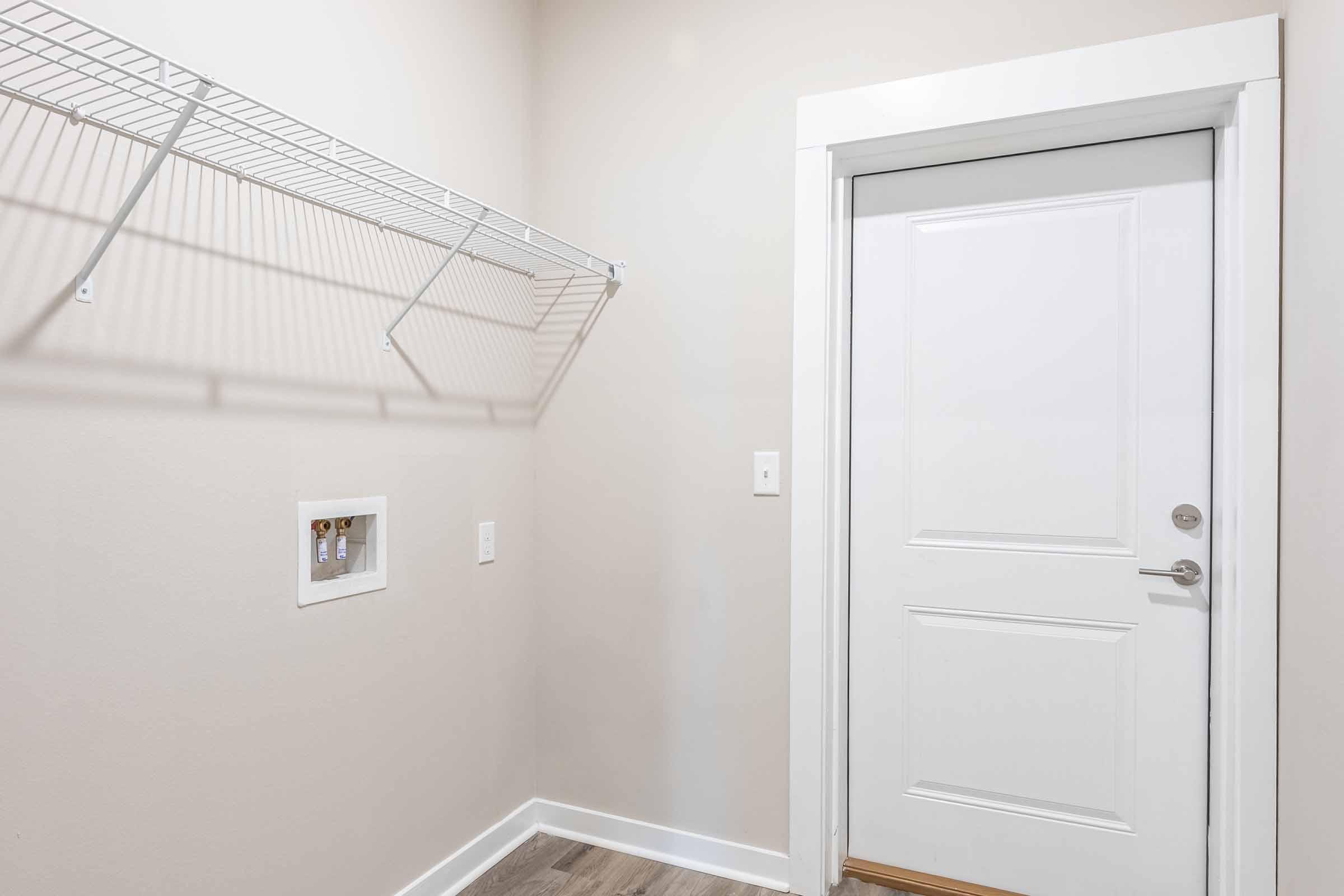 A blank wall with a white door and a simple wall-mounted wire shelf above. There is a small electrical outlet and a light switch on the wall next to the door. The floor has light-colored wood-like flooring, creating a clean and minimalistic look in the space.