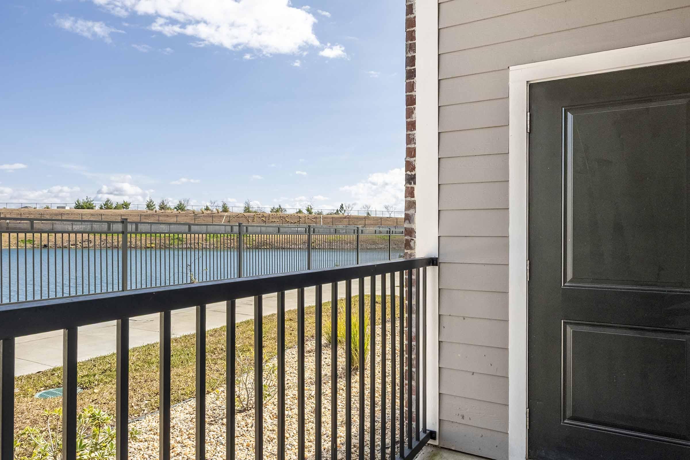 View of a balcony with a black door leading to an interior space. The balcony features a metal railing overlooking a calm body of water and a landscaped grassy area. In the background, a low wall and trees line the opposite bank under a blue sky with scattered clouds.