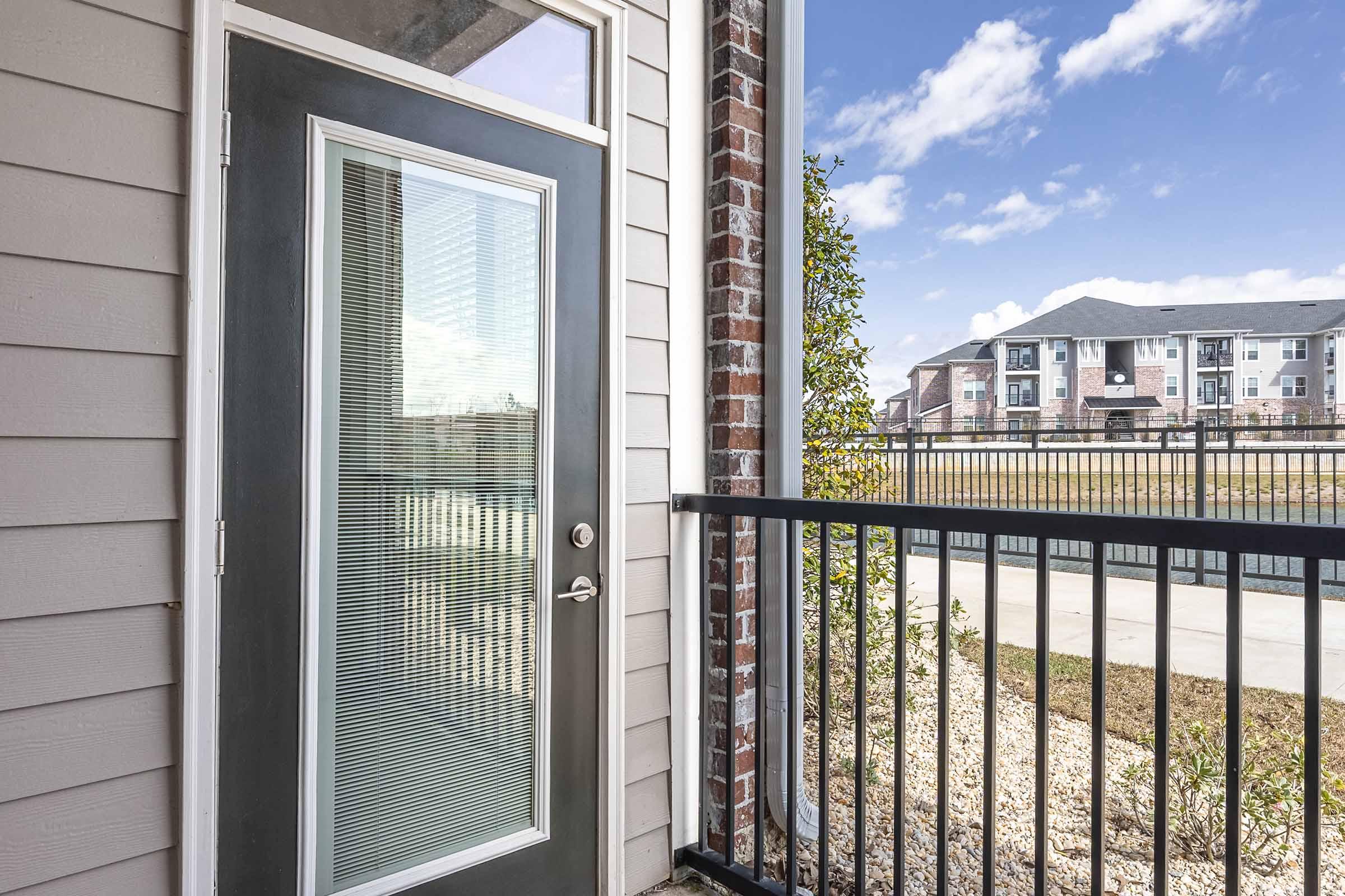 A close-up view of a modern apartment entry door with a glass panel, located on a balcony. The exterior features gray siding and brick accents. In the background, there are landscaped areas and a view of nearby apartment buildings under a blue sky with scattered clouds.