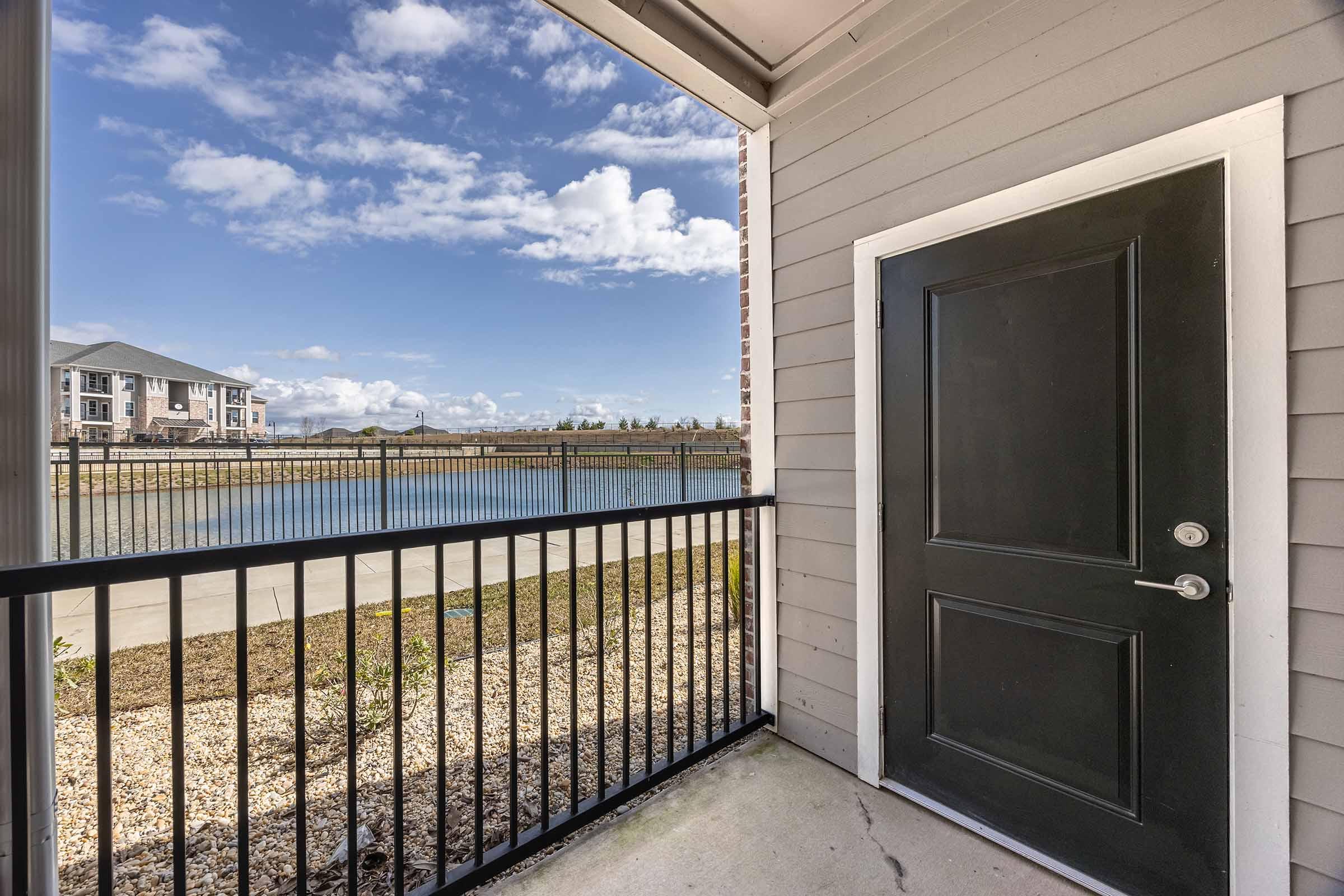 View from an apartment entrance, featuring a black door and a balcony overlooking a calm lake. In the background, green grass and a fence border the water, with buildings visible on the opposite shore under a partly cloudy sky.