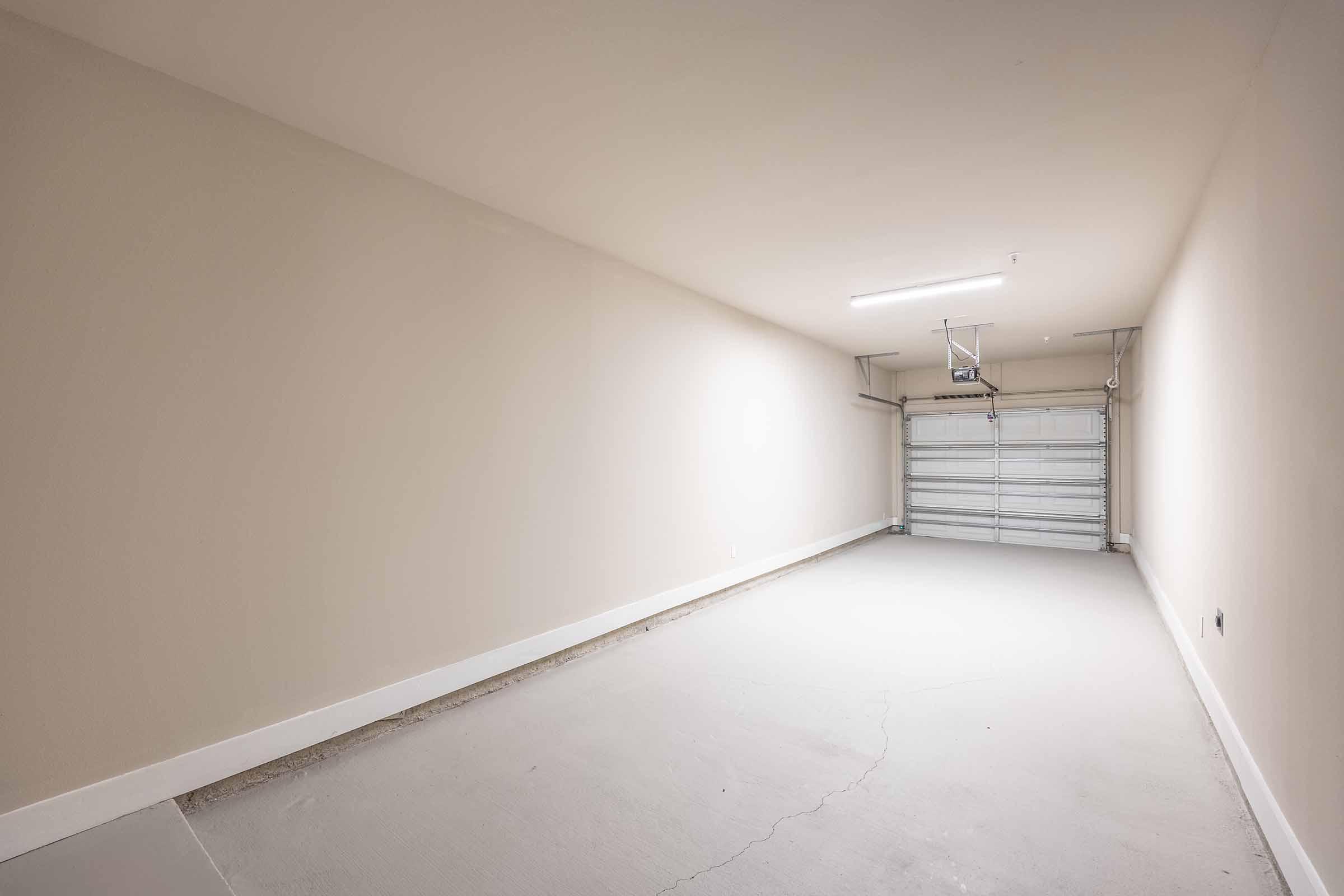 Empty garage space with plain walls, a concrete floor, and a closed garage door. There is overhead lighting but no furniture or items present, creating a minimalist environment. The overall color scheme is neutral and simplistic.