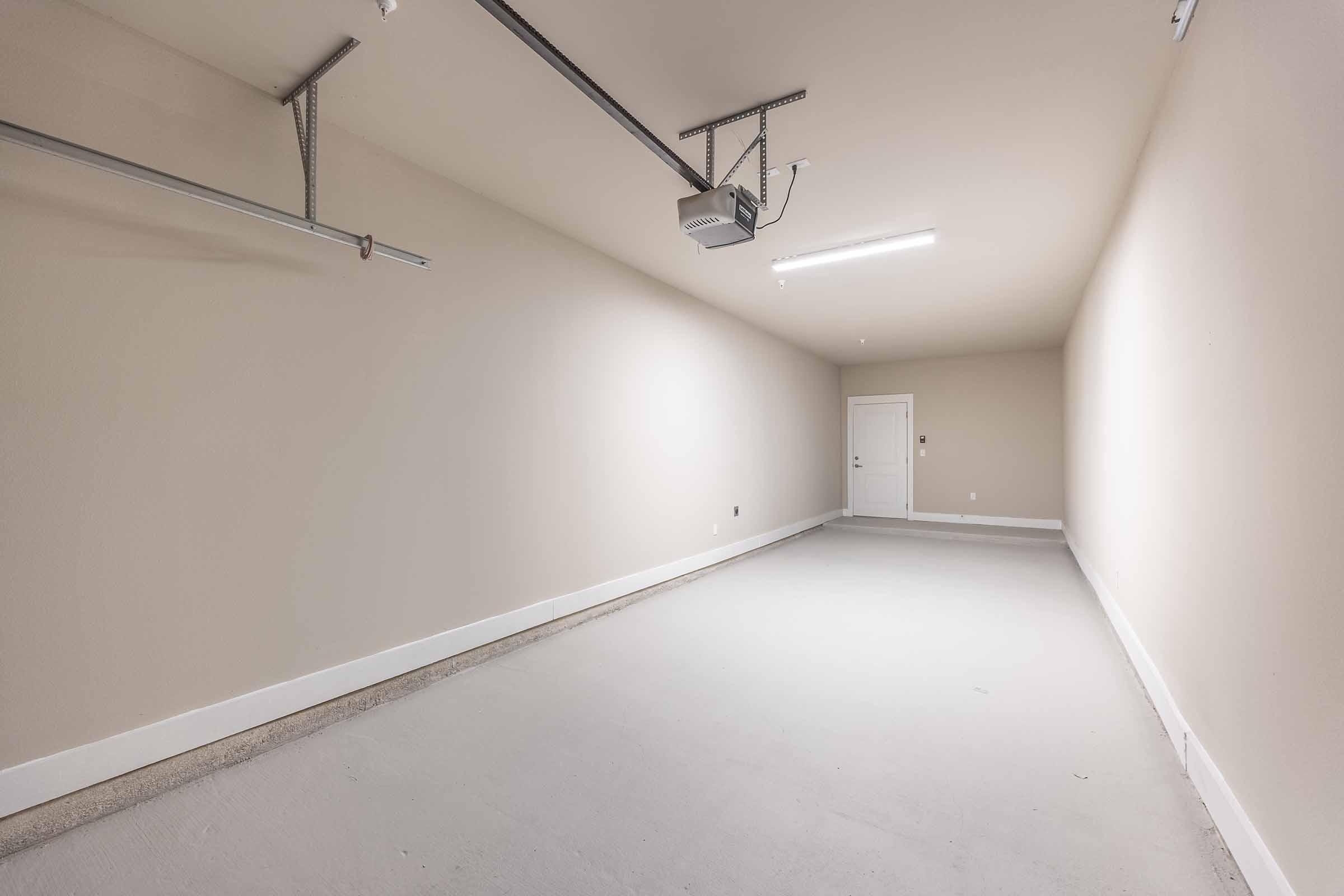 A spacious and empty garage with beige walls and a concrete floor. The ceiling features a garage door opener, and there is a white door visible at the far end, leading to another area. The lighting is bright, creating a clean and uncluttered atmosphere.