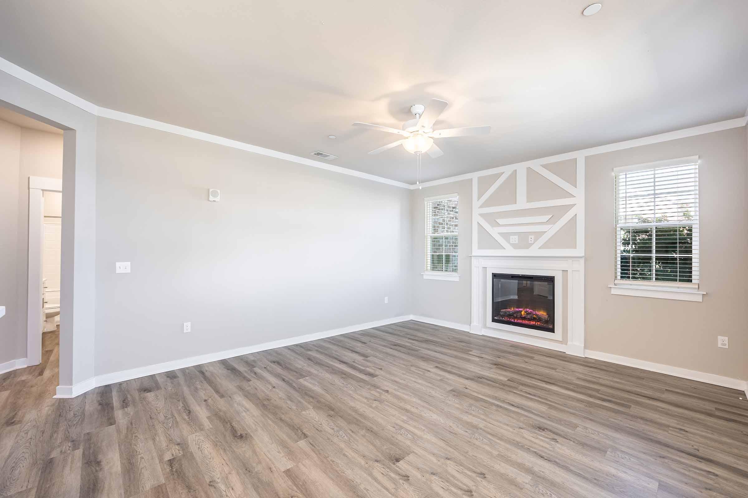 A spacious, empty living room with light-colored walls and a ceiling fan. It features a fireplace with a decorative mantel, hardwood-like flooring, and large windows that allow natural light. The layout includes an opening to another room, enhancing the sense of openness.
