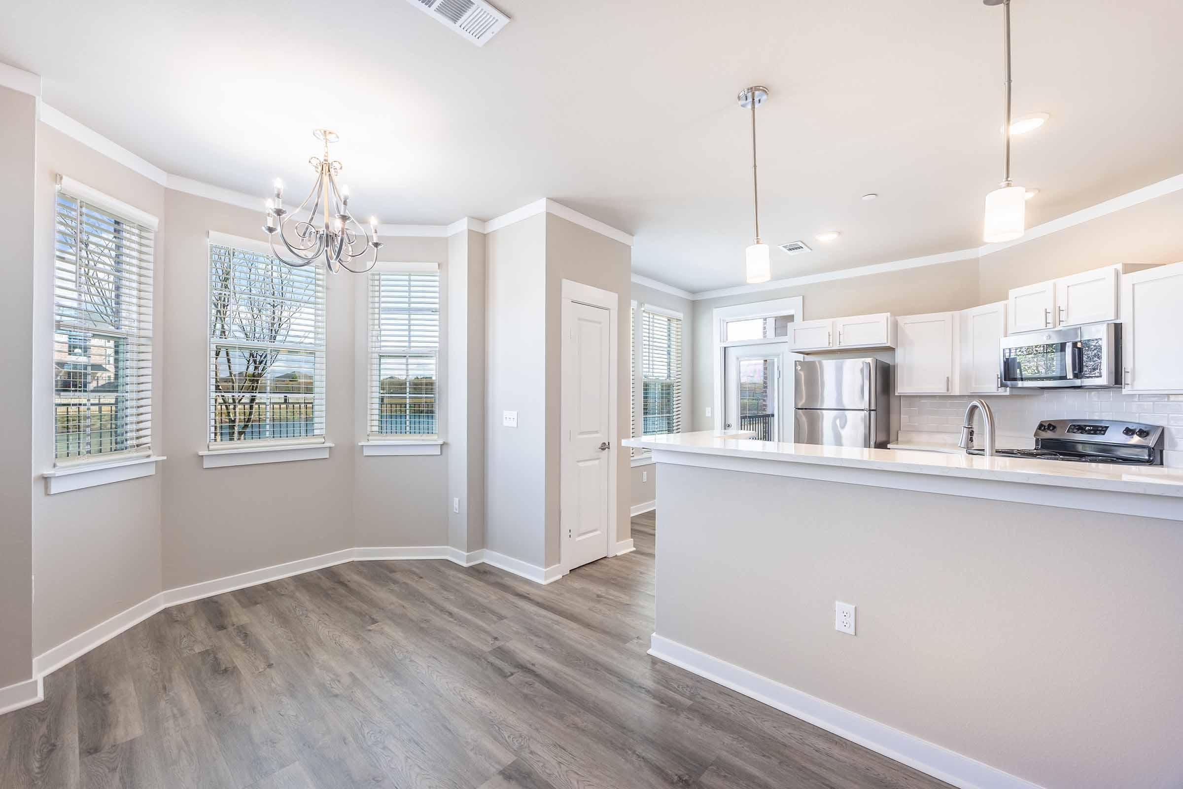Bright, modern kitchen area with white cabinets and stainless steel appliances. Features include a sleek countertop, overhead lighting, and a cozy dining space near large windows with horizontal blinds. The room has light-colored walls and laminate flooring, creating a spacious and airy atmosphere.