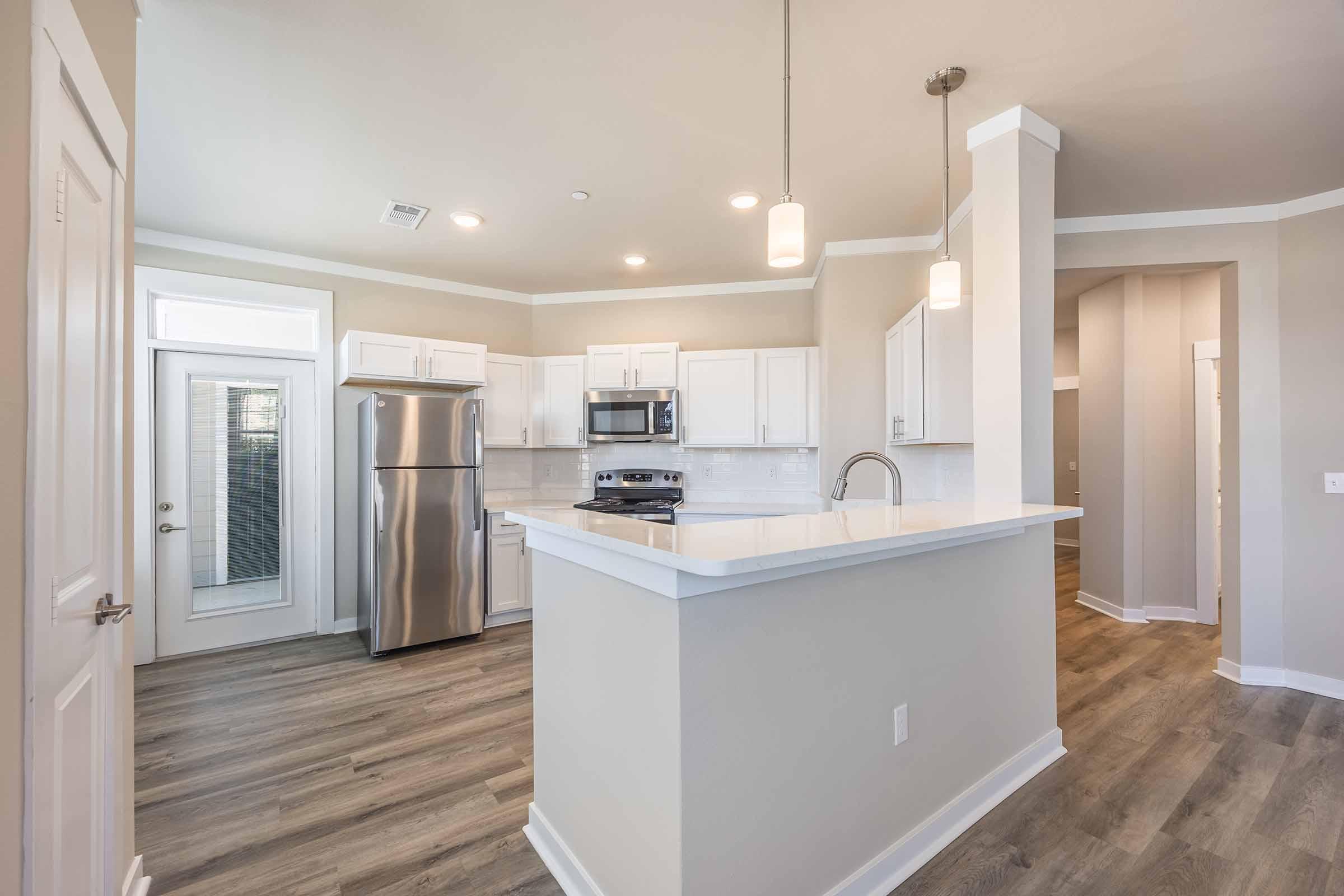 Modern kitchen with stainless steel appliances including a refrigerator and microwave. Light gray walls and cabinets create a fresh look, complemented by a white countertop. Pendant lights hang above the counter, and there is a door leading outside, giving the space an open and inviting feel.