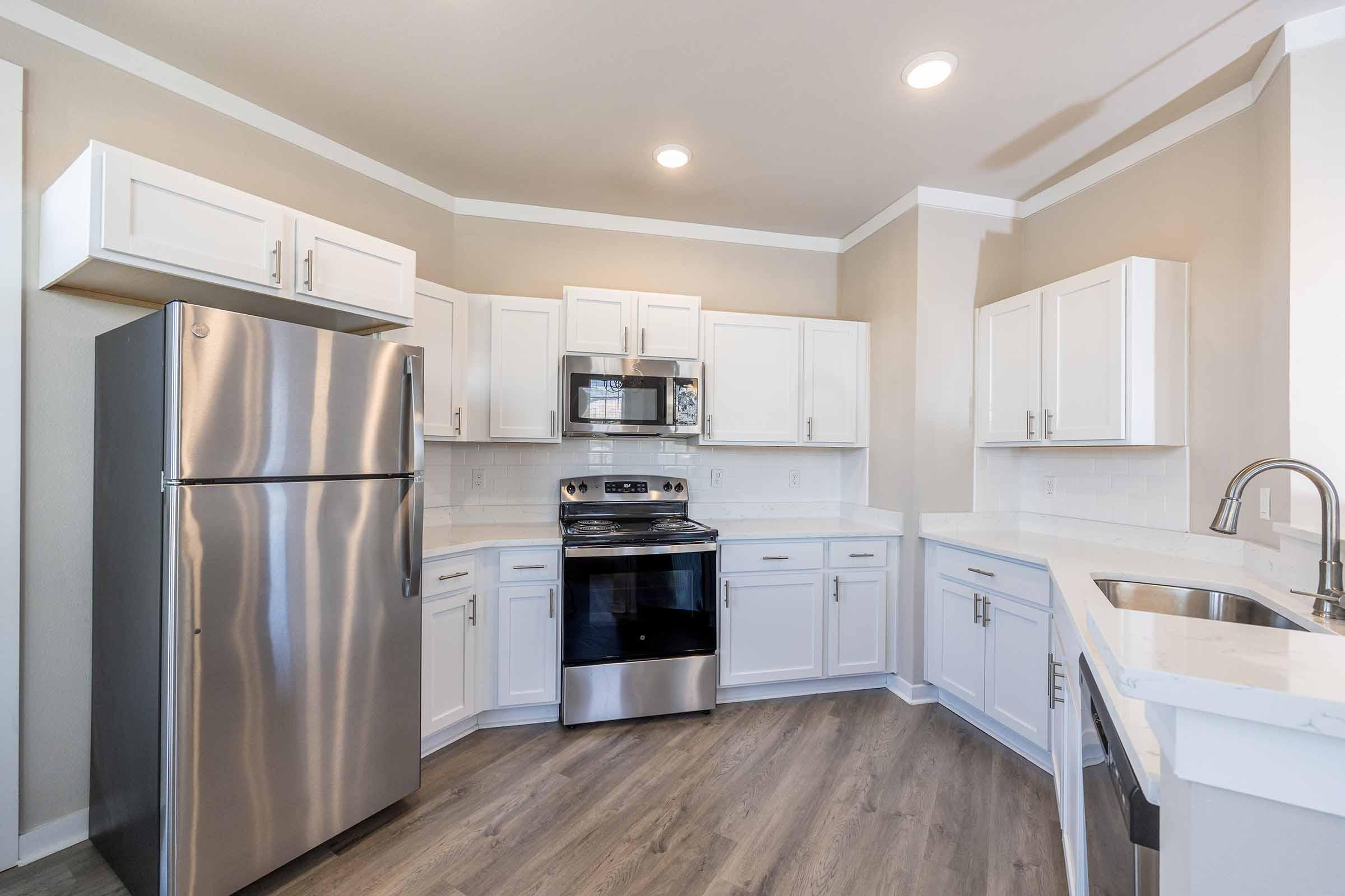Modern kitchen featuring stainless steel appliances, including a refrigerator, stove, and microwave. White cabinetry complements a light countertop. The space has a neutral color scheme with recessed lighting and a spacious layout, enhancing its functionality and aesthetics.