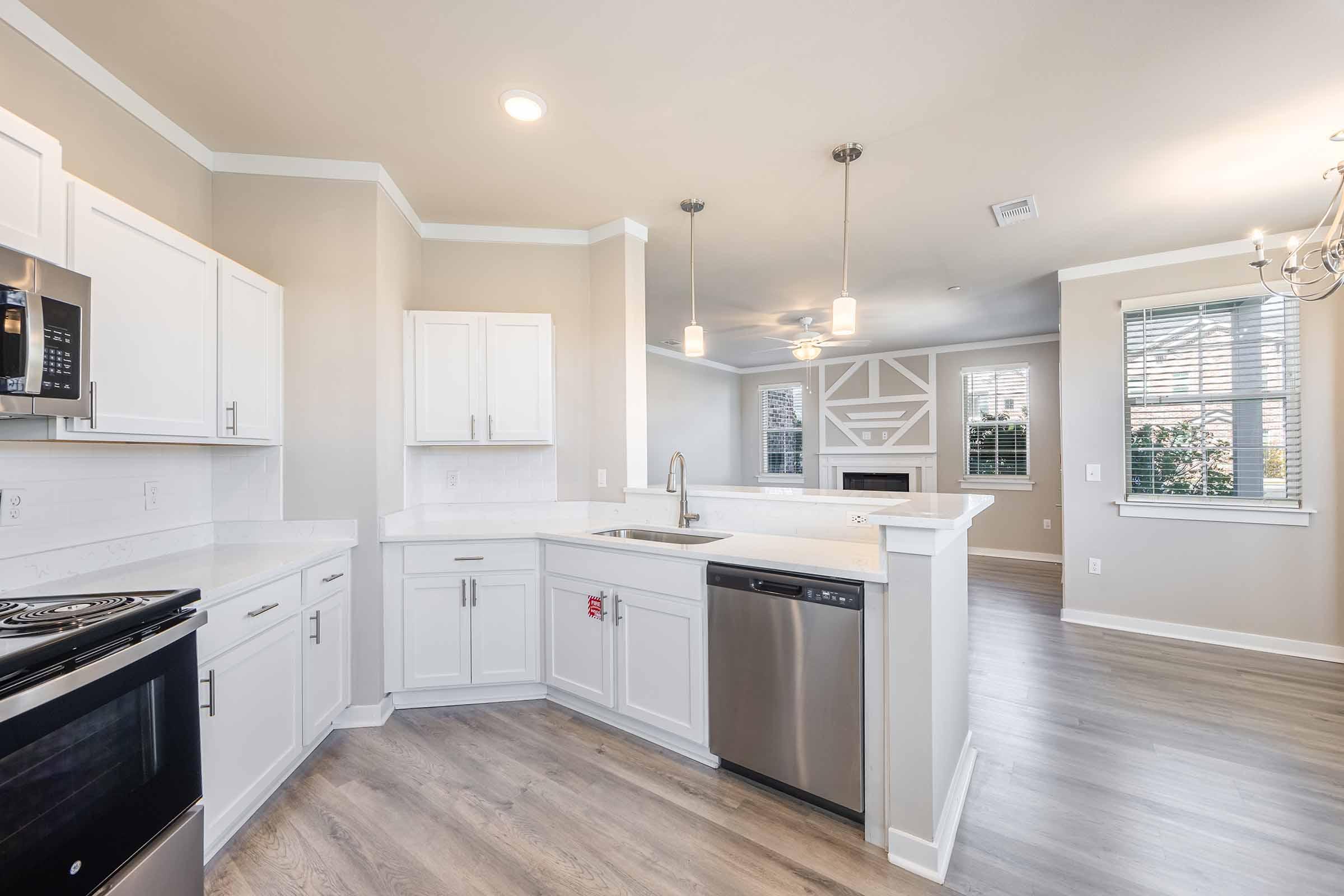 Modern kitchen with white cabinetry, stainless steel appliances, and a large sink. Features an open layout that connects to a bright living area with large windows. The flooring is light-colored wood, and there are pendant lights hanging above the island, creating a contemporary and inviting space.