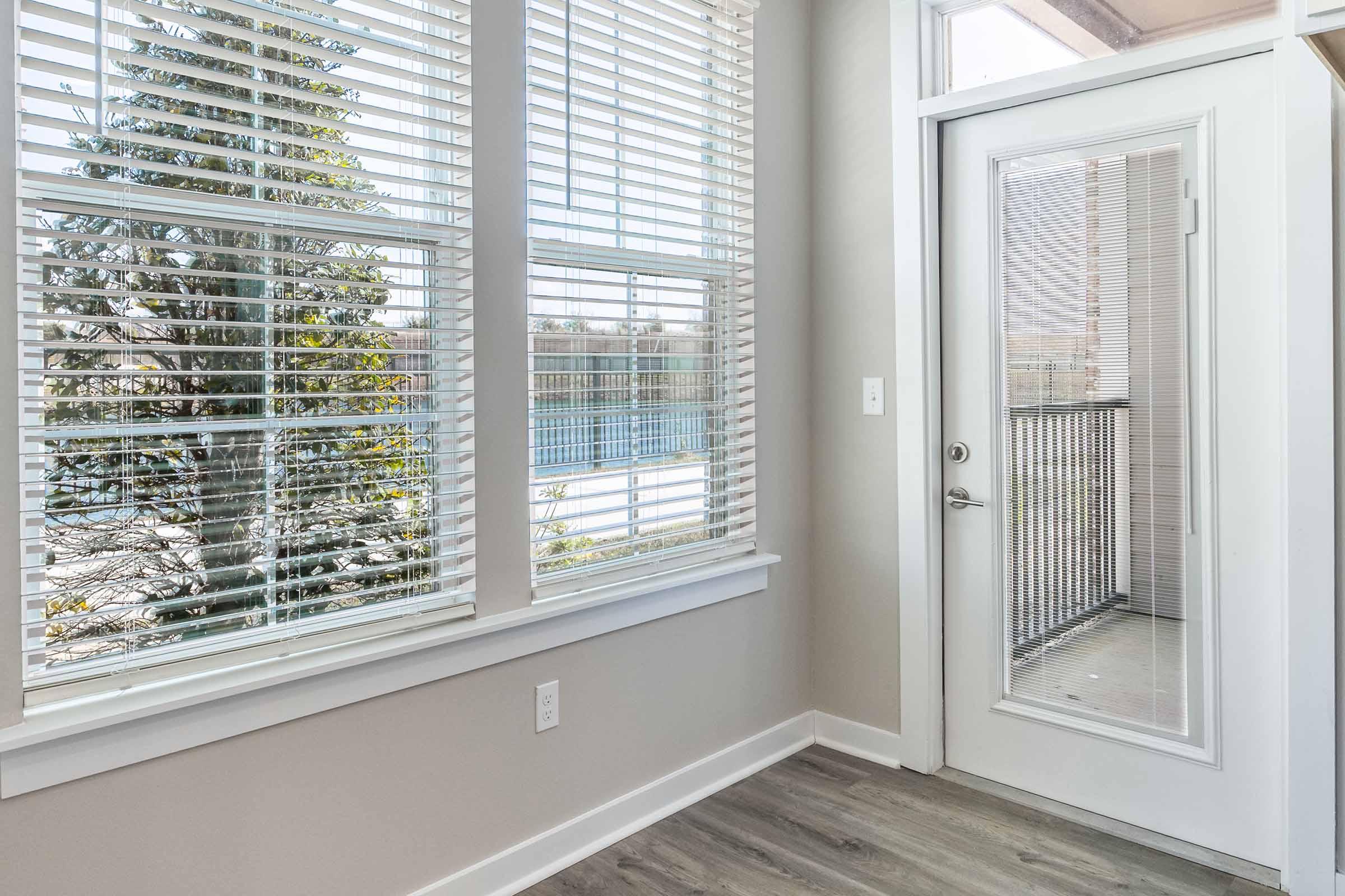 A bright room with large windows featuring white blinds, offering a view of trees outside. There is a glass door leading to a balcony, and the flooring is light-colored wood. The walls are painted in a soft neutral shade, creating a calm and inviting atmosphere.