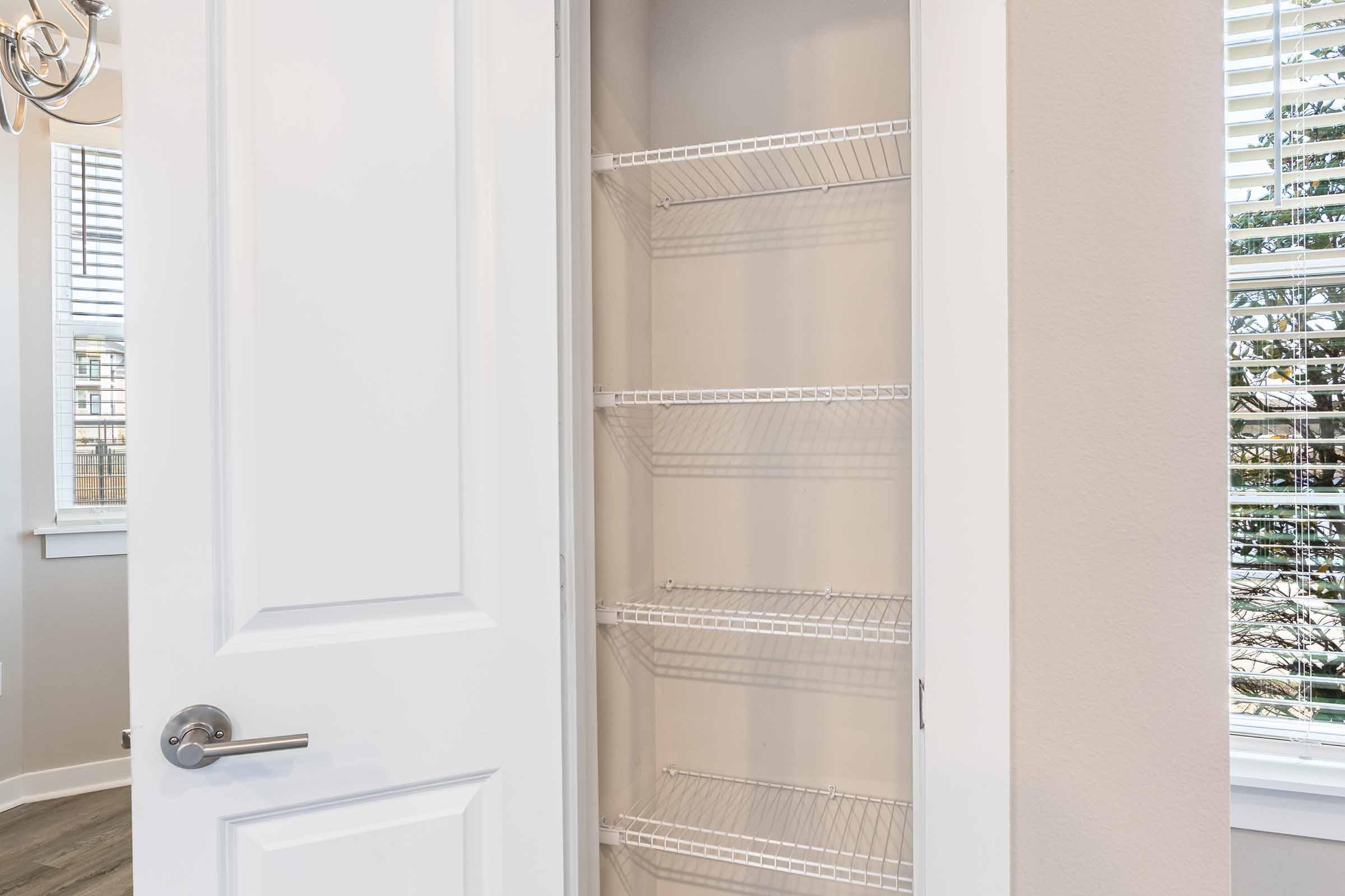 A clean, empty closet with white wire shelves and a light beige wall. The closet door is slightly open, showing the shelves inside. Natural light filters in through nearby windows, illuminating the space.