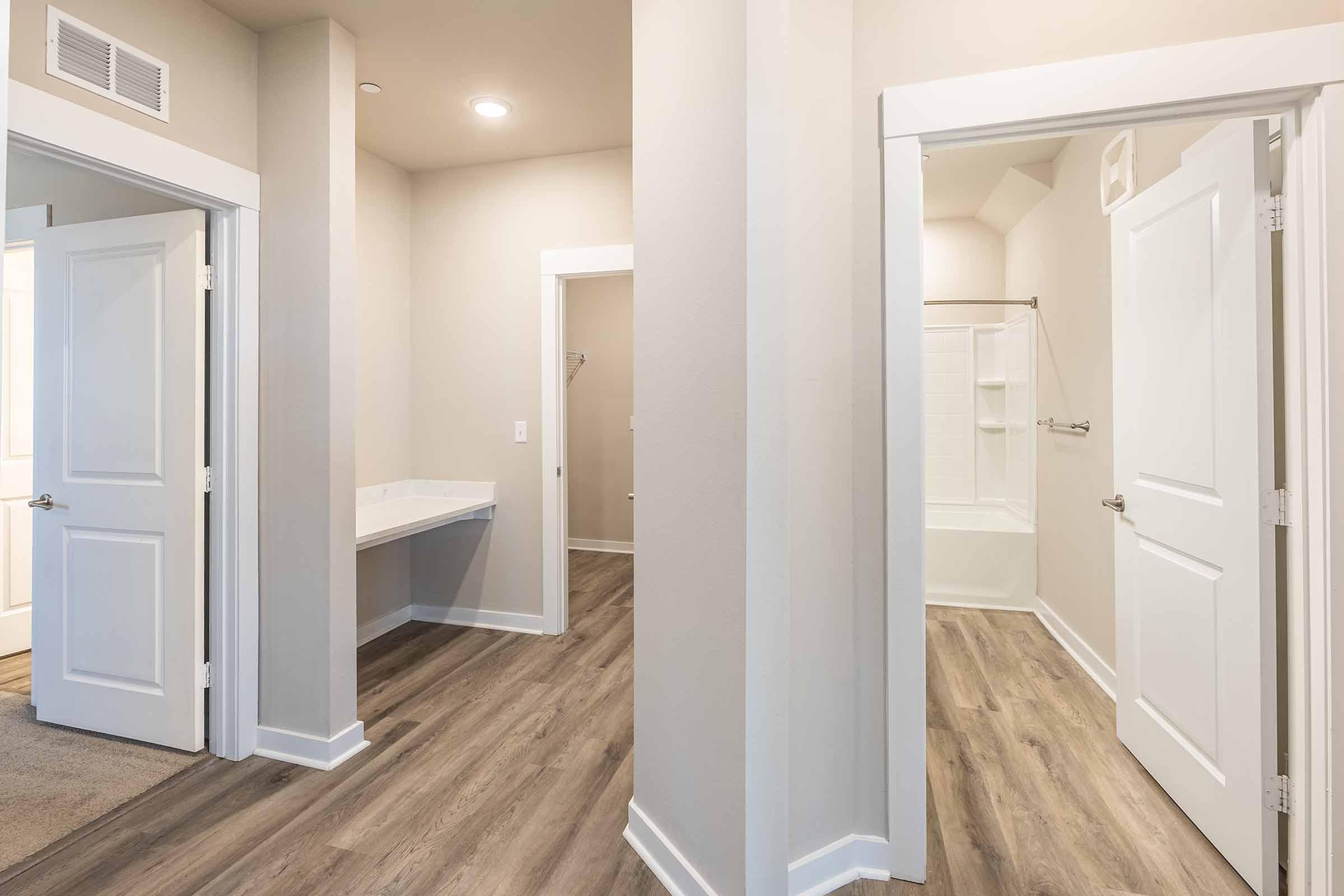 Interior view of a modern bathroom with a white bathtub and shower, open doorways leading to adjacent rooms, and light-colored walls. The flooring is a wood-like laminate. The space is well-lit with ceiling lights and a welcoming layout.