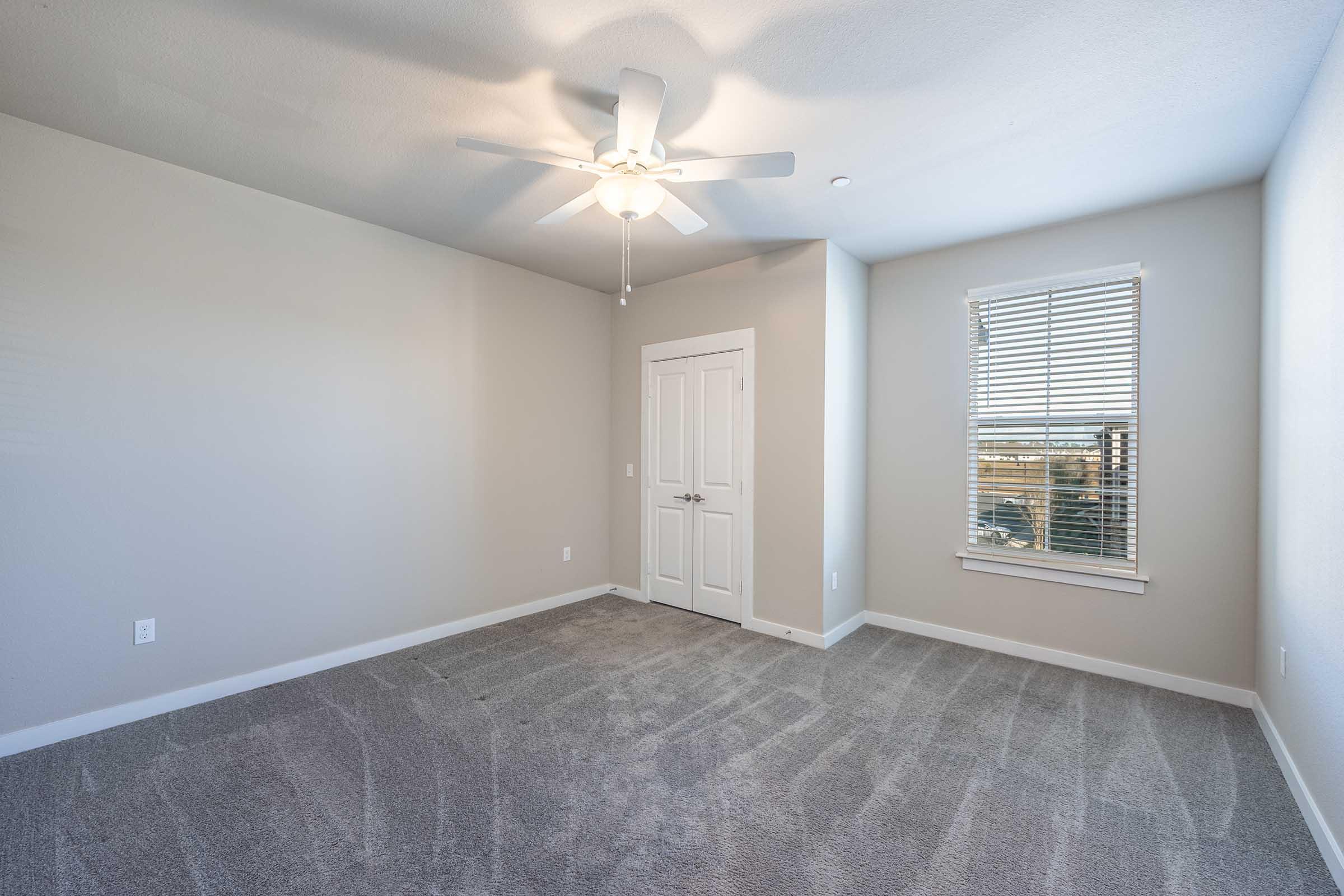 A well-lit, empty room with light gray walls and plush carpet. Features a ceiling fan, a closed door, and a window with blinds allowing natural light to enter. The room has a modern and clean aesthetic, making it suitable for various uses.