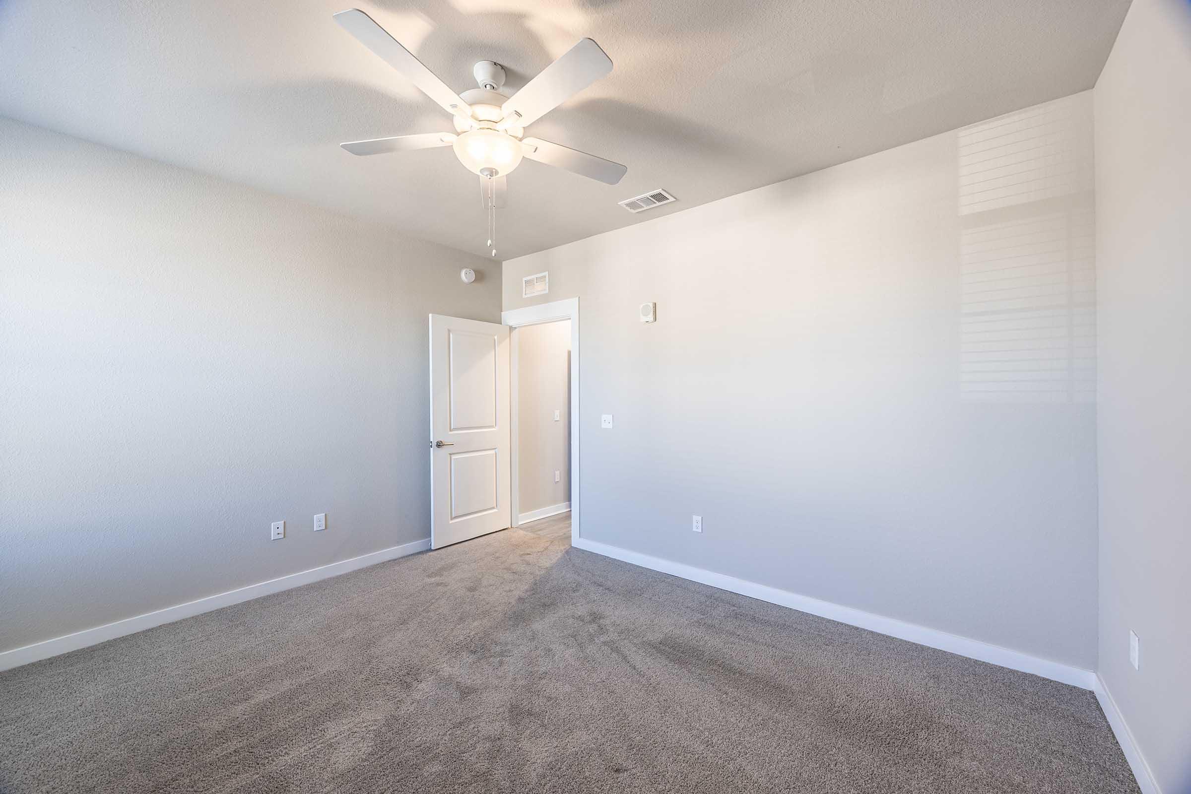 Empty room with light gray walls and carpet, featuring a ceiling fan. A door on the left leads to another room, while the opposite wall has a light switch and no furniture, creating a spacious and bright atmosphere. Natural light is visible from a nearby window.
