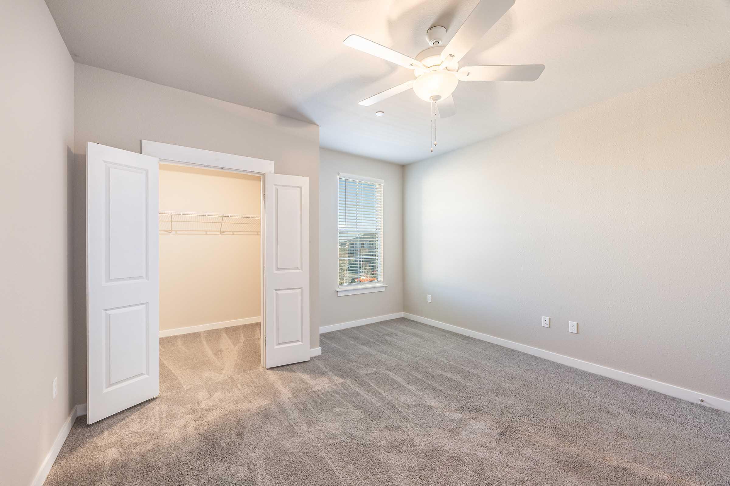 A spacious, empty bedroom featuring light gray walls and carpeted flooring. The room includes a ceiling fan, a window with blinds allowing natural light, and a closet with bi-fold doors open, displaying a simple wire shelf inside.