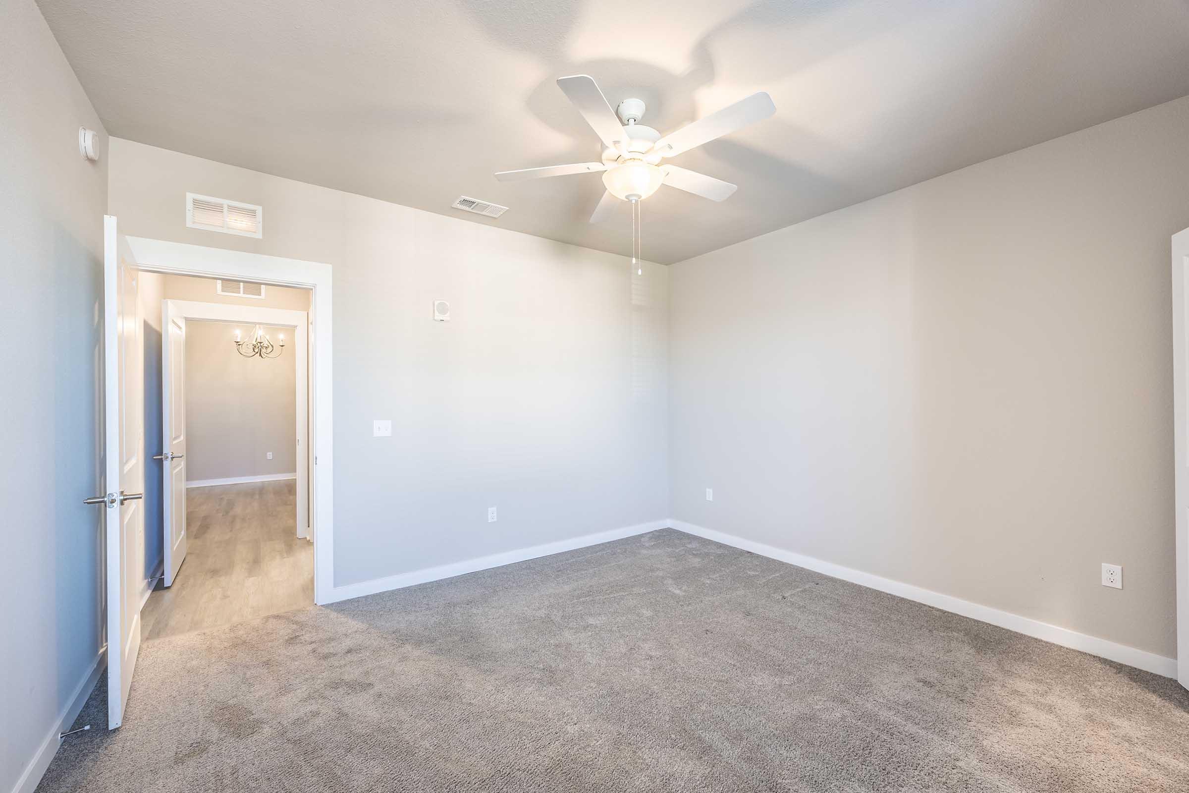 A well-lit, empty room with light gray walls and carpet. A ceiling fan is installed, and there is a doorway on the left leading to a hallway. The light from the door illuminates the space, creating a welcoming atmosphere.