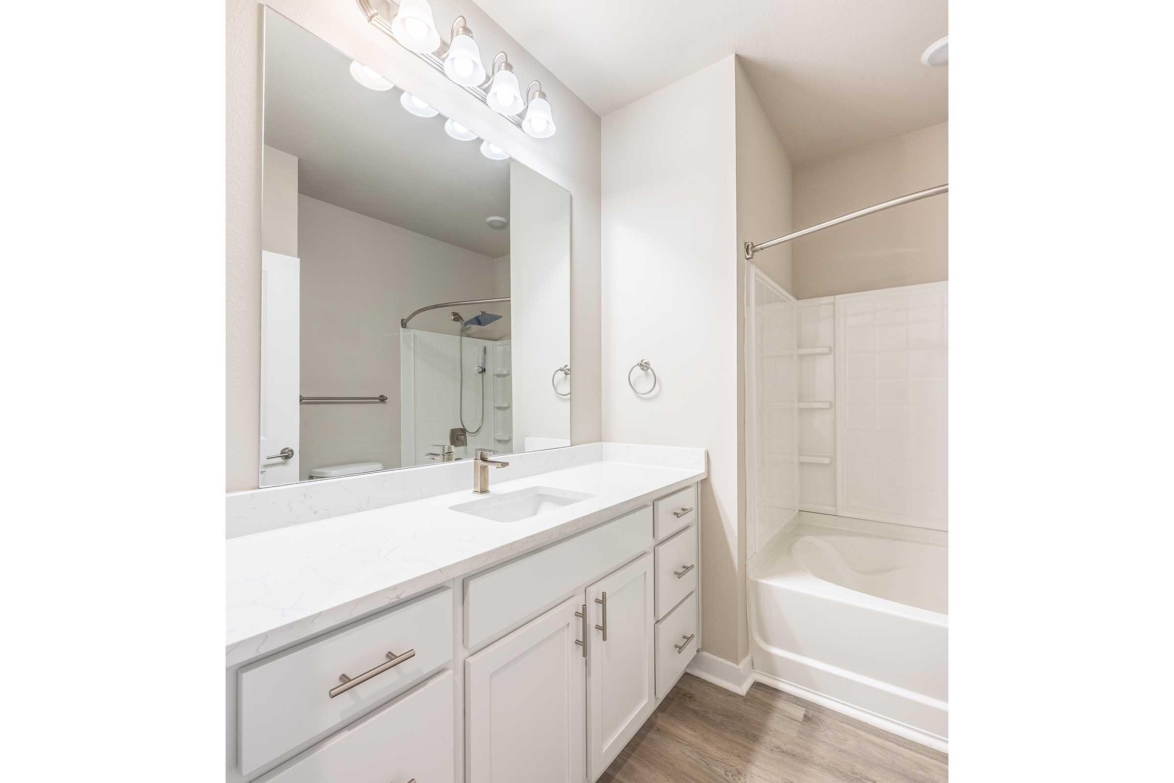 A modern bathroom featuring a double vanity with white cabinetry, a sleek sink, and a large mirror above. The space includes a bathtub/shower combo with white tile surround and a light fixture with five bulbs. The walls are painted in a neutral color, and the flooring is a light wood finish.