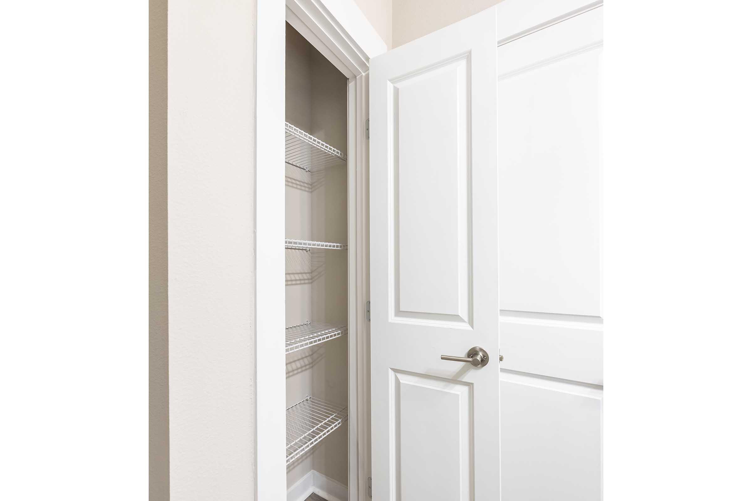 A narrow closet with an open white door, featuring several wire shelves inside. The walls are painted a light color, and the floor is not visible. The door has a silver handle and there is a sense of clean, minimalist design.