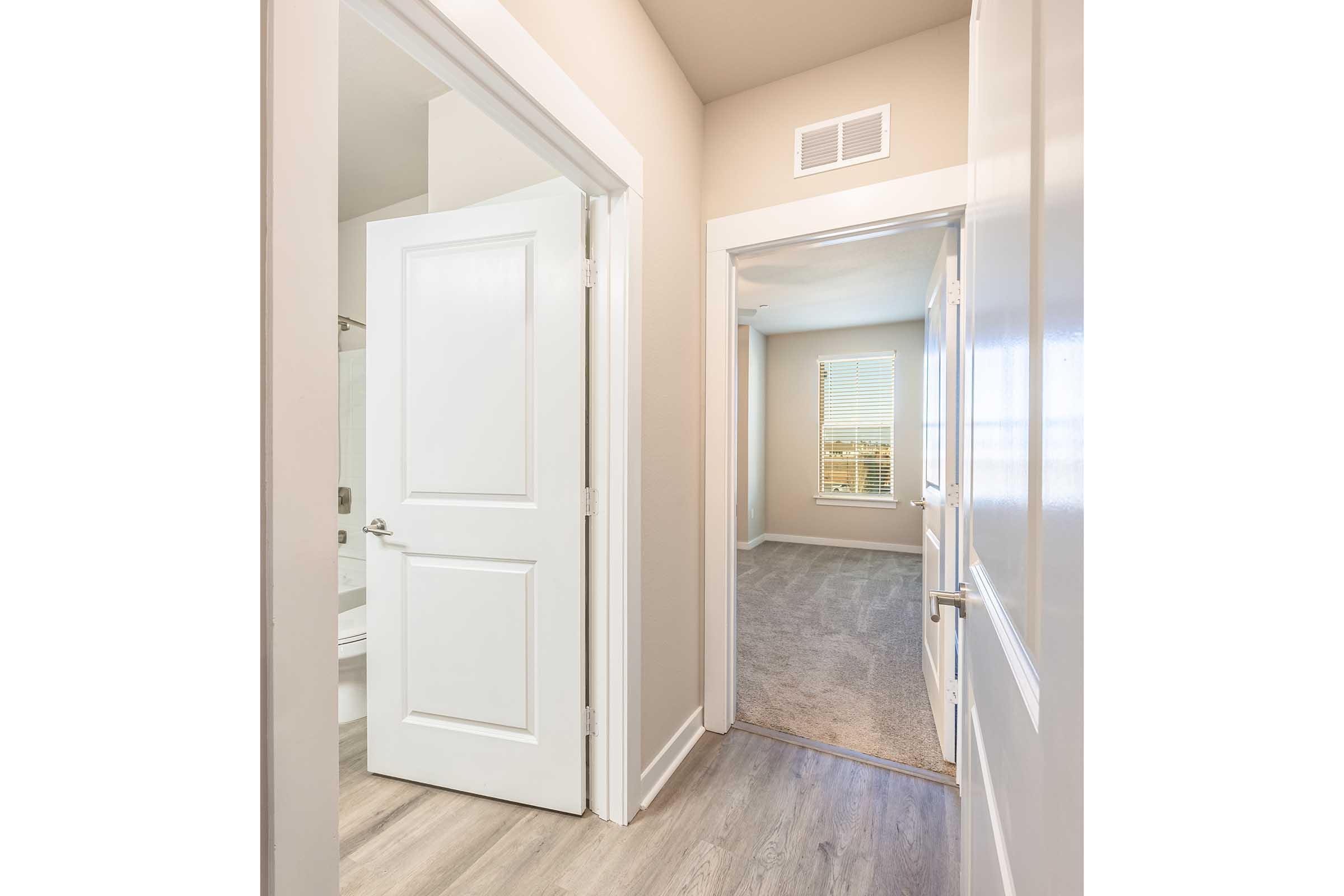 A hallway view featuring two white doors, one leading to a bathroom and the other to a room with a window. The walls are painted in a light color, and the flooring is a light wood finish. Natural light fills the space, creating a bright and airy atmosphere.