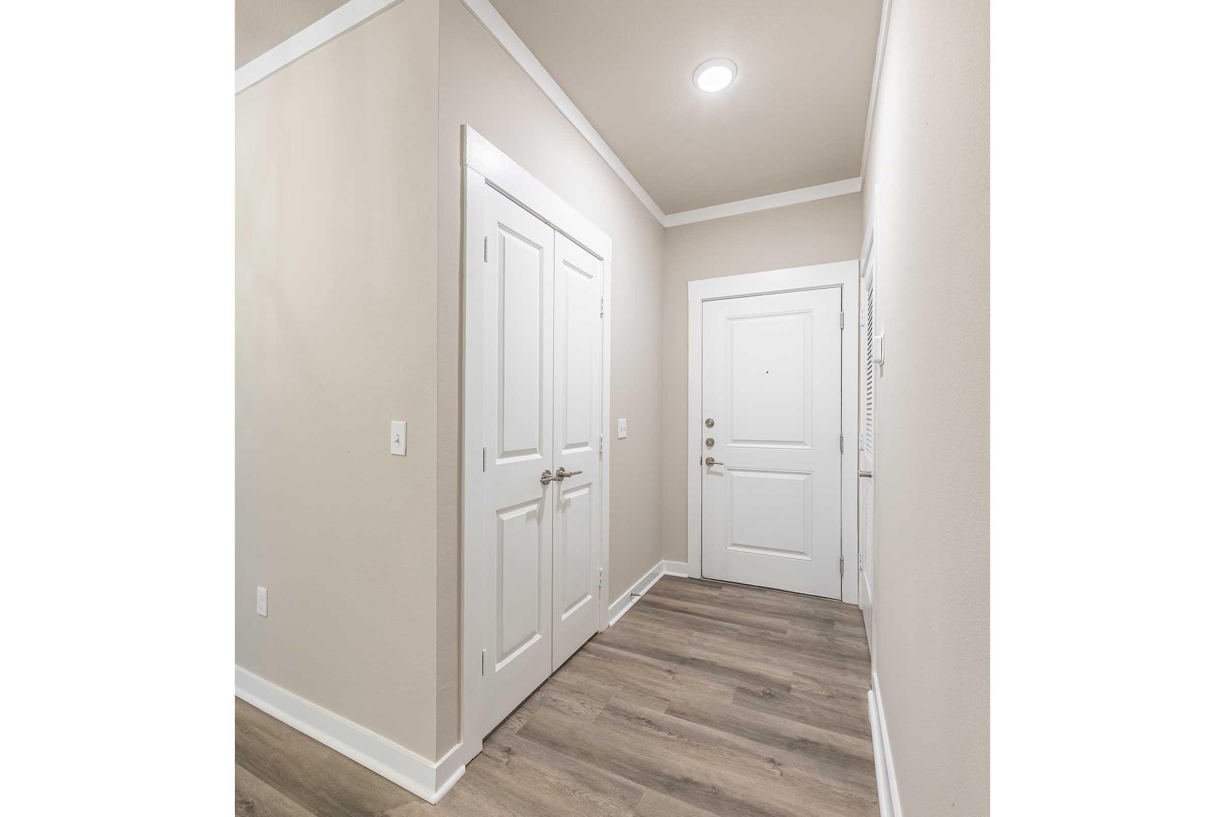 A well-lit hallway featuring beige walls, light hardwood flooring, and white trim. There are two closed doors on the left and a main entrance door on the right. The space is modern and inviting, with a simple, minimalist design.