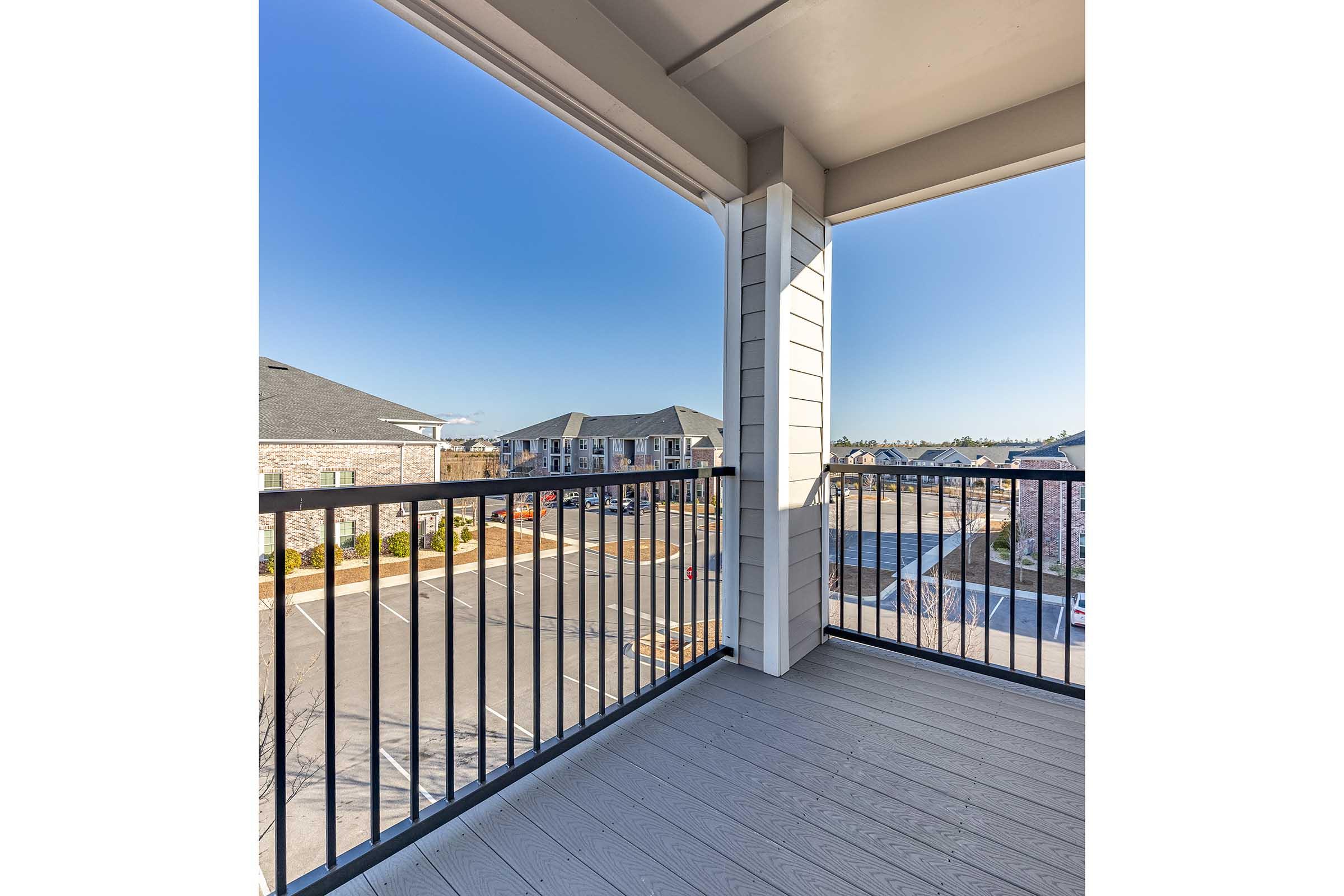 View from a balcony featuring a railing, overlooking a residential area with multiple buildings in the background. The sky is clear and blue, indicating a sunny day. The foreground showcases wooden decking, suggesting a modern apartment or condo setting.