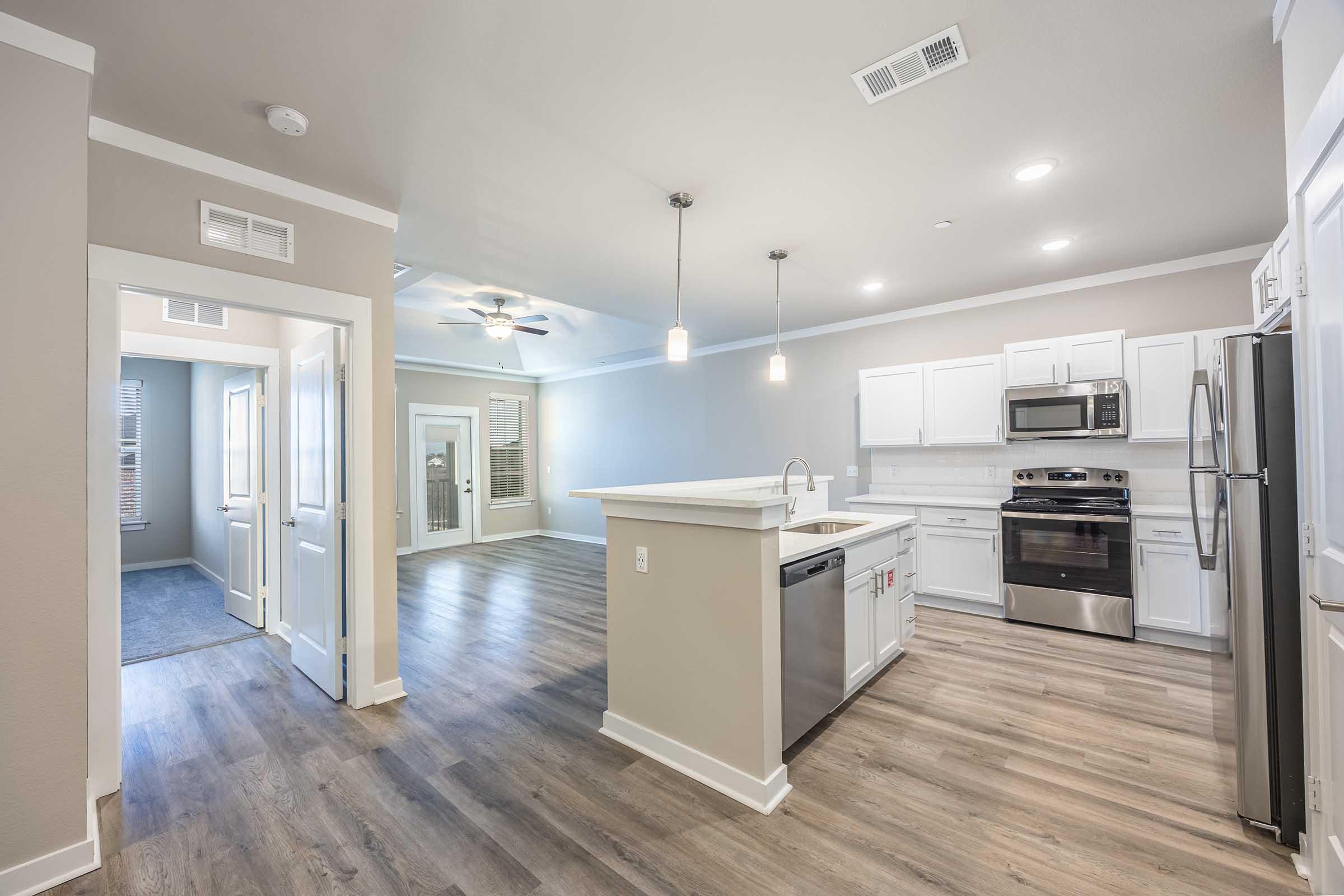 A modern kitchen with white cabinets, stainless steel appliances, and a breakfast bar, connected to a light-filled living area. The flooring is a wood-like laminate. To the left, a doorway leads to another room, while large windows allow natural light to enter, creating a spacious and inviting atmosphere.