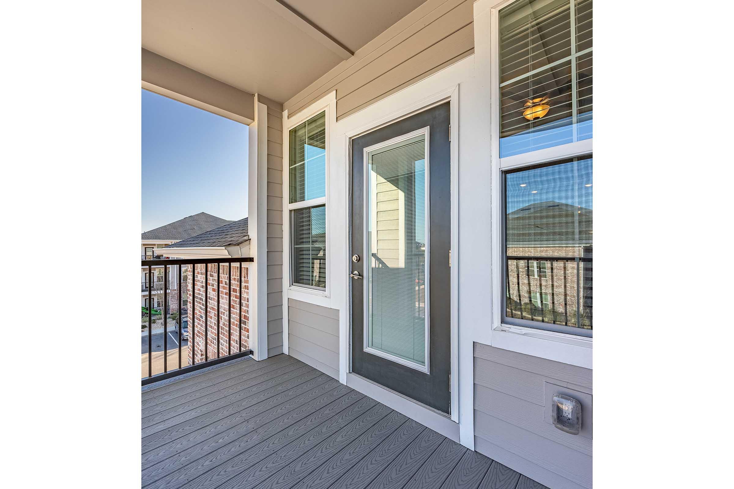 A view of a modern balcony featuring a sliding glass door with a dark frame, large windows providing natural light, and a wooden deck floor. The surrounding area shows a distant neighborhood landscape under clear skies.