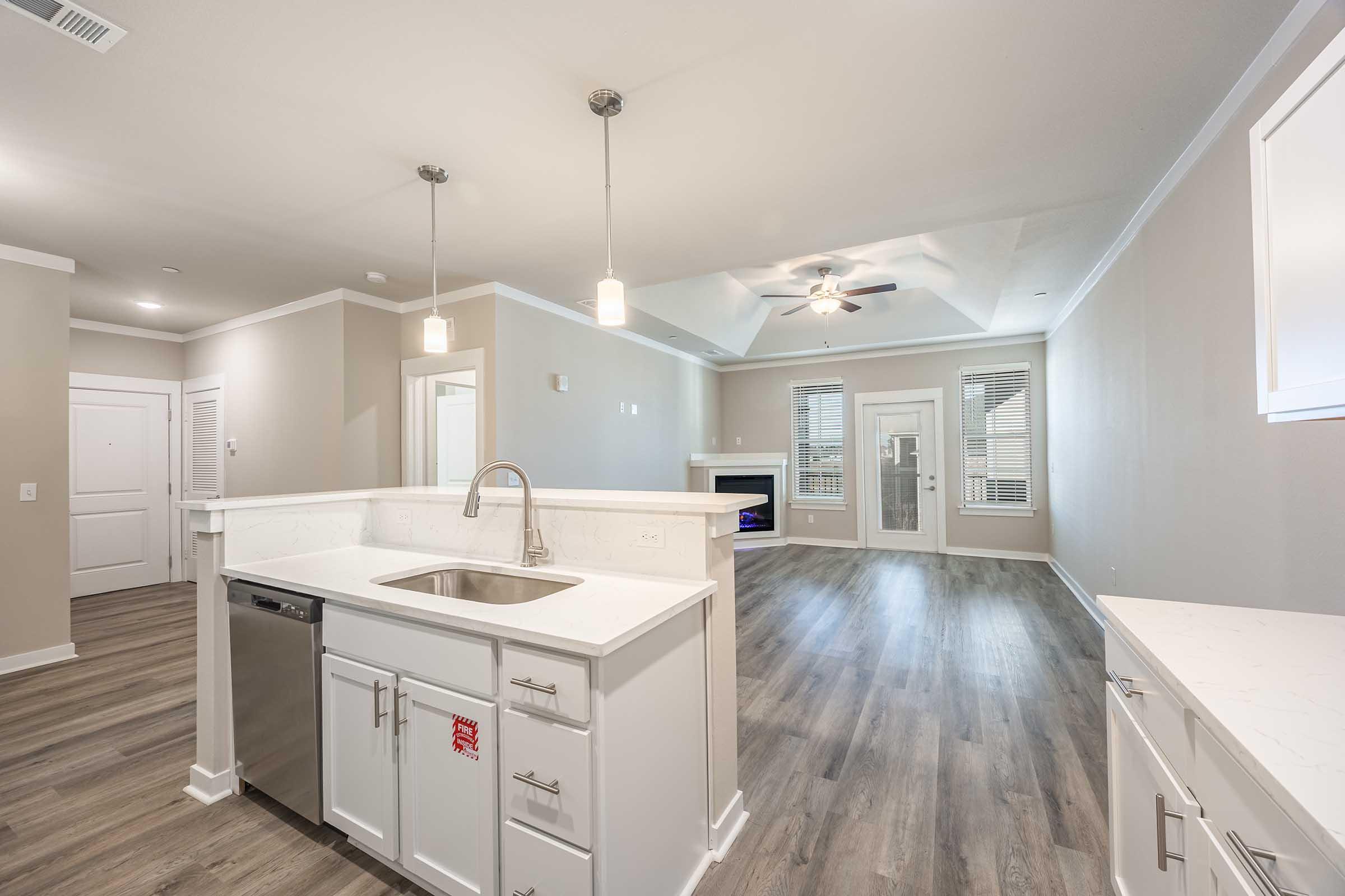A modern kitchen with a white countertop and stainless steel sink, overlooking a spacious living area. The room features light-colored walls, wood-like flooring, recessed lighting, and large windows allowing natural light. A ceiling fan is visible, and there is a doorway leading to another room.