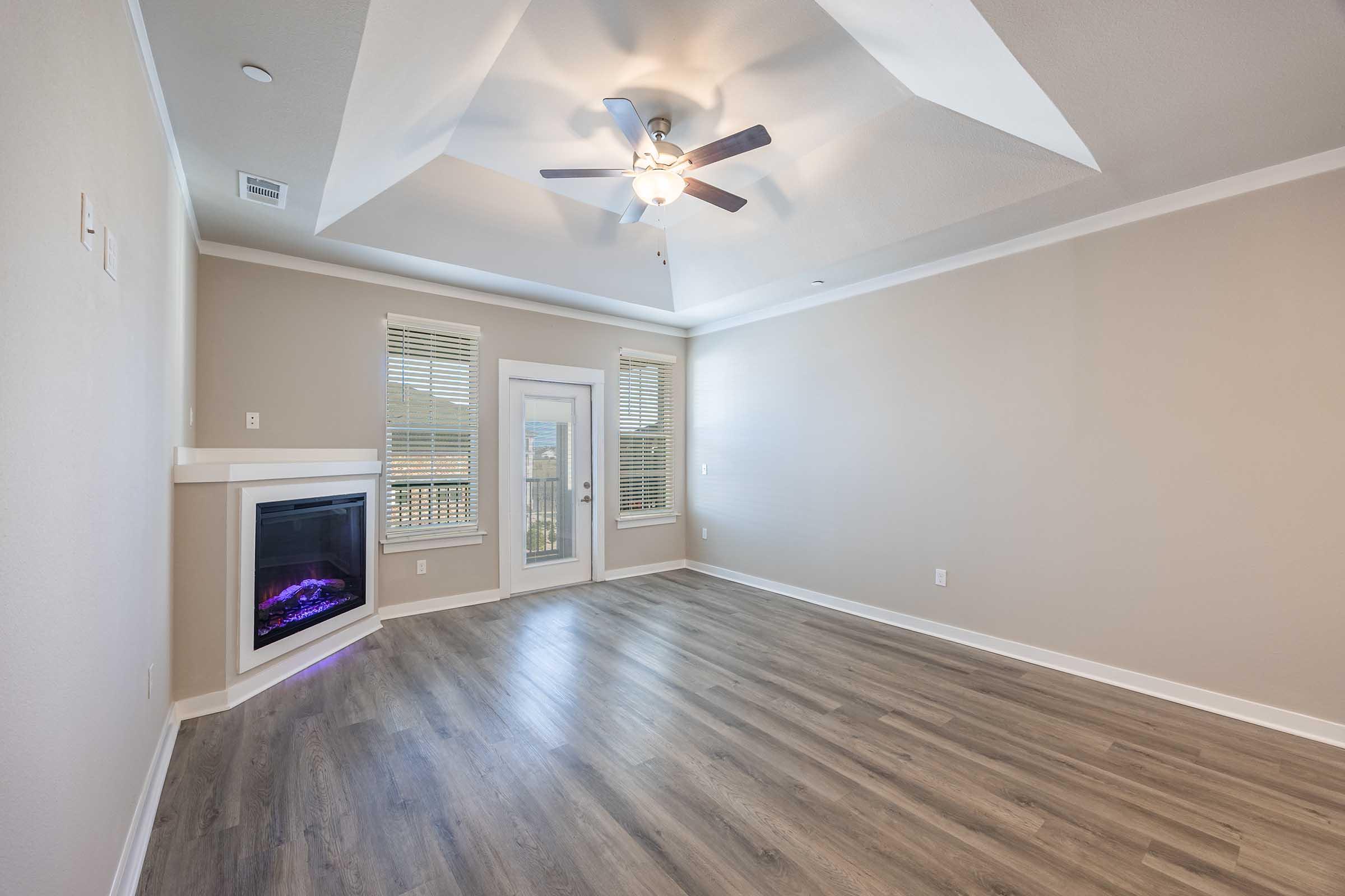 A spacious living room with a modern ceiling fan, large windows, and a built-in electric fireplace. The walls are painted in a light color, and the flooring is a warm-toned wood laminate. Natural light streams in, enhancing the airy feel of the room.