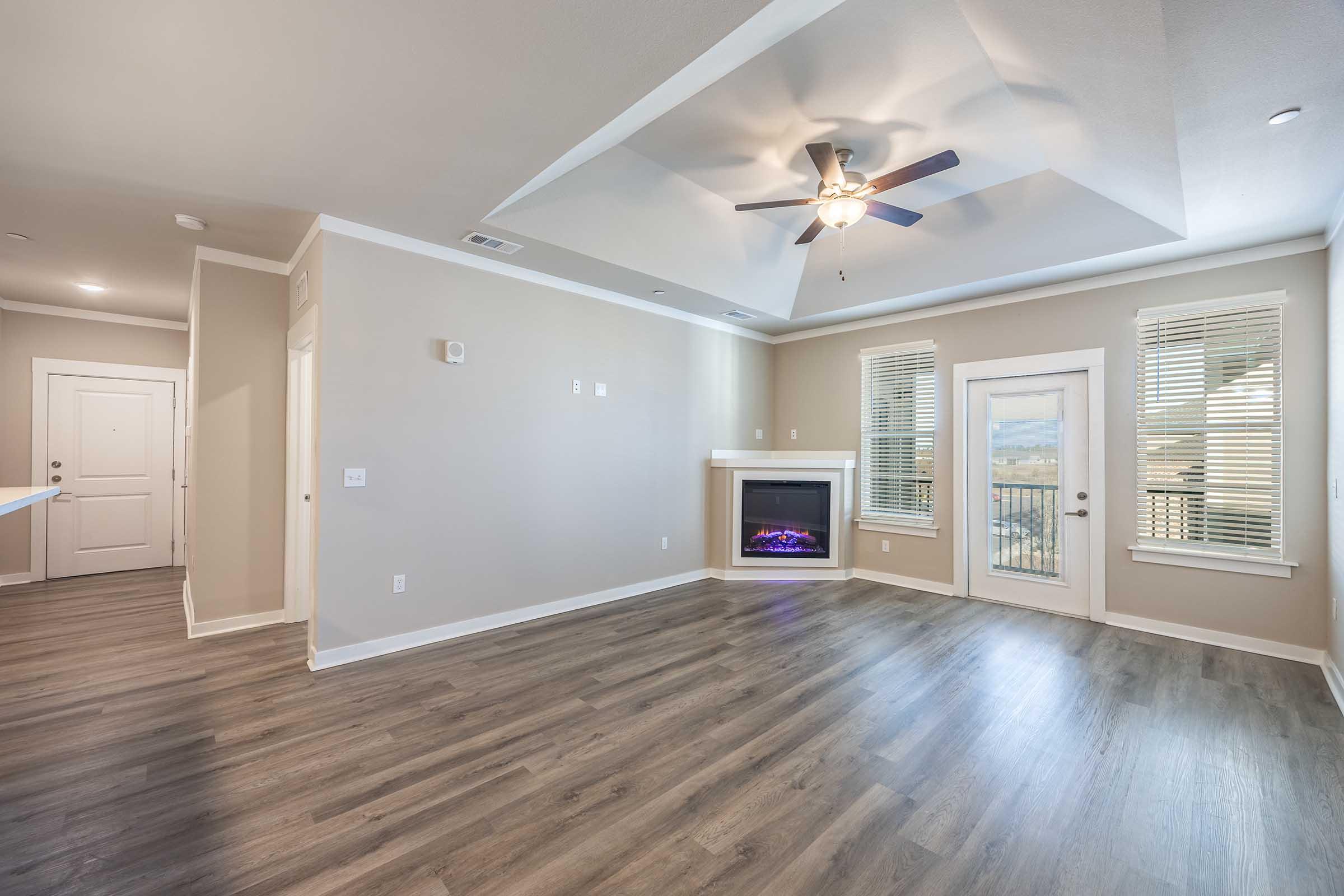 A spacious, well-lit living room featuring a ceiling fan, modern light fixtures, and a cozy electric fireplace. Large windows allow natural light to fill the space, and the flooring is hardwood. The room has neutral-colored walls and access to a balcony through a glass door.