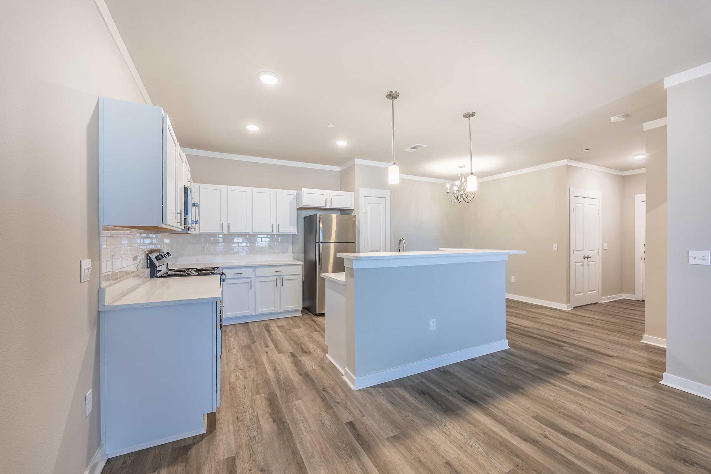 A modern, well-lit kitchen with white cabinetry, stainless steel appliances, and a large island. The floors are a light wood grain, and the walls are a soft beige. A dining area with a chandelier is visible, along with an entryway in the background.