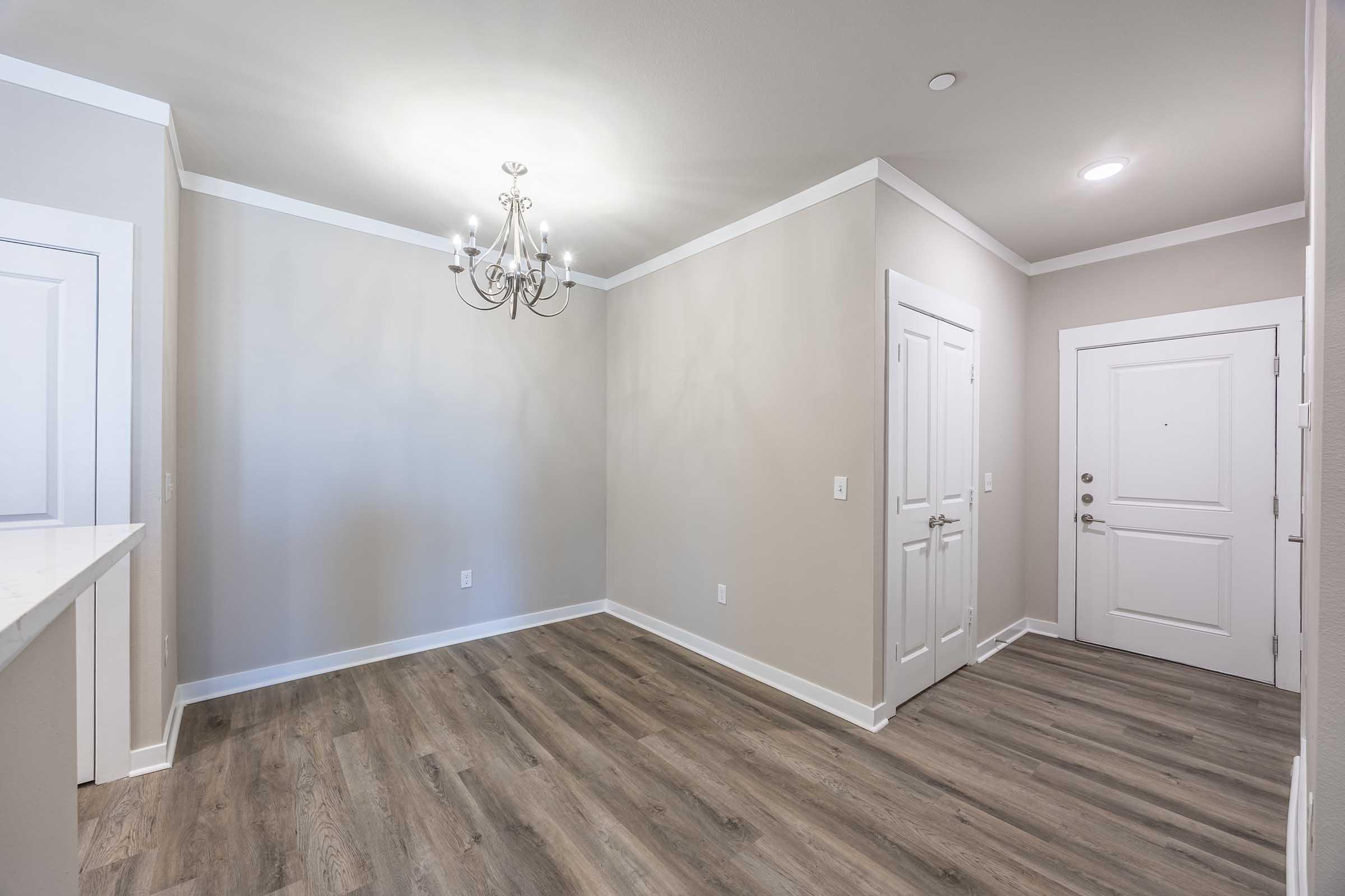 A well-lit entryway featuring light gray walls, a modern chandelier, and hardwood-style flooring. The space includes a door to the left and a white panel door to the right. There is a small ledge or counter near the entrance, and the room is empty, creating a welcoming atmosphere for inhabitants.