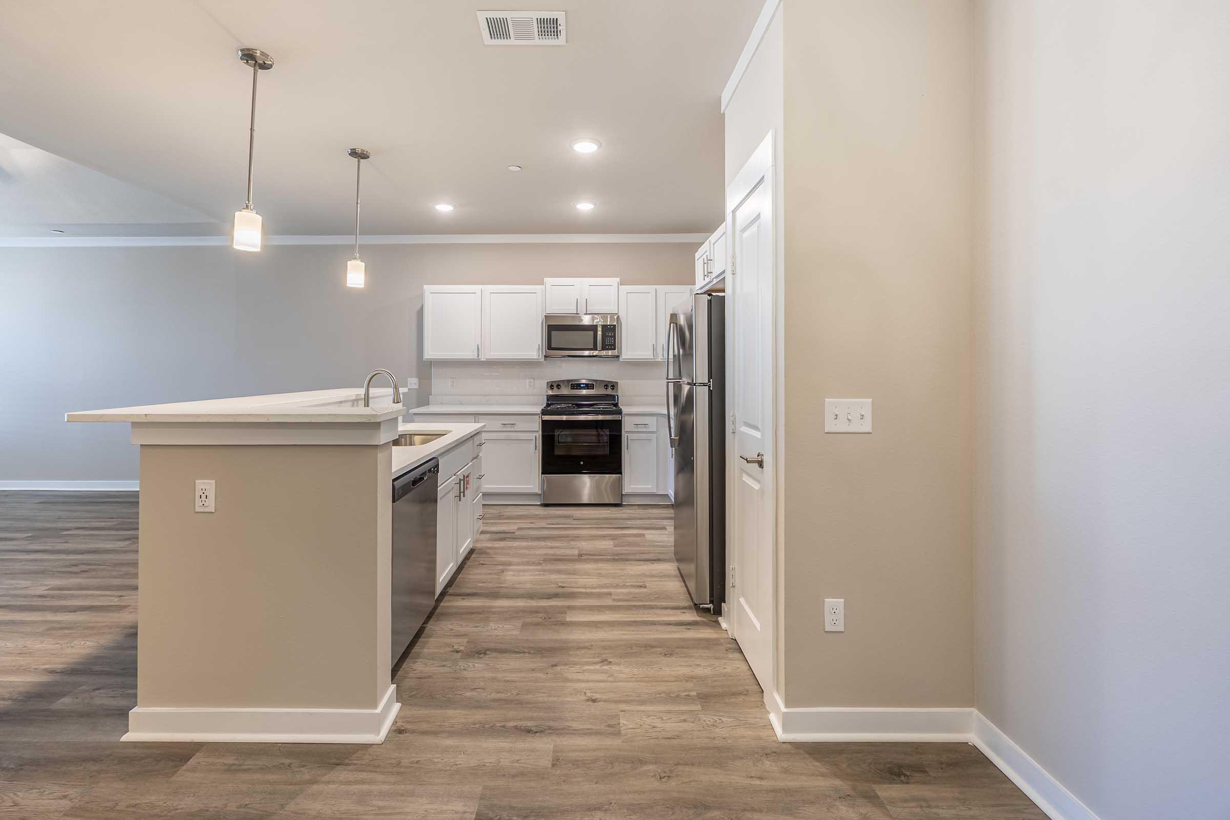A modern kitchen featuring a spacious layout with an island counter, stainless steel appliances, white cabinetry, and light-colored walls. The flooring is a warm brown wood-like texture, creating a contemporary and inviting atmosphere. Lighting fixtures hang gently from the ceiling.