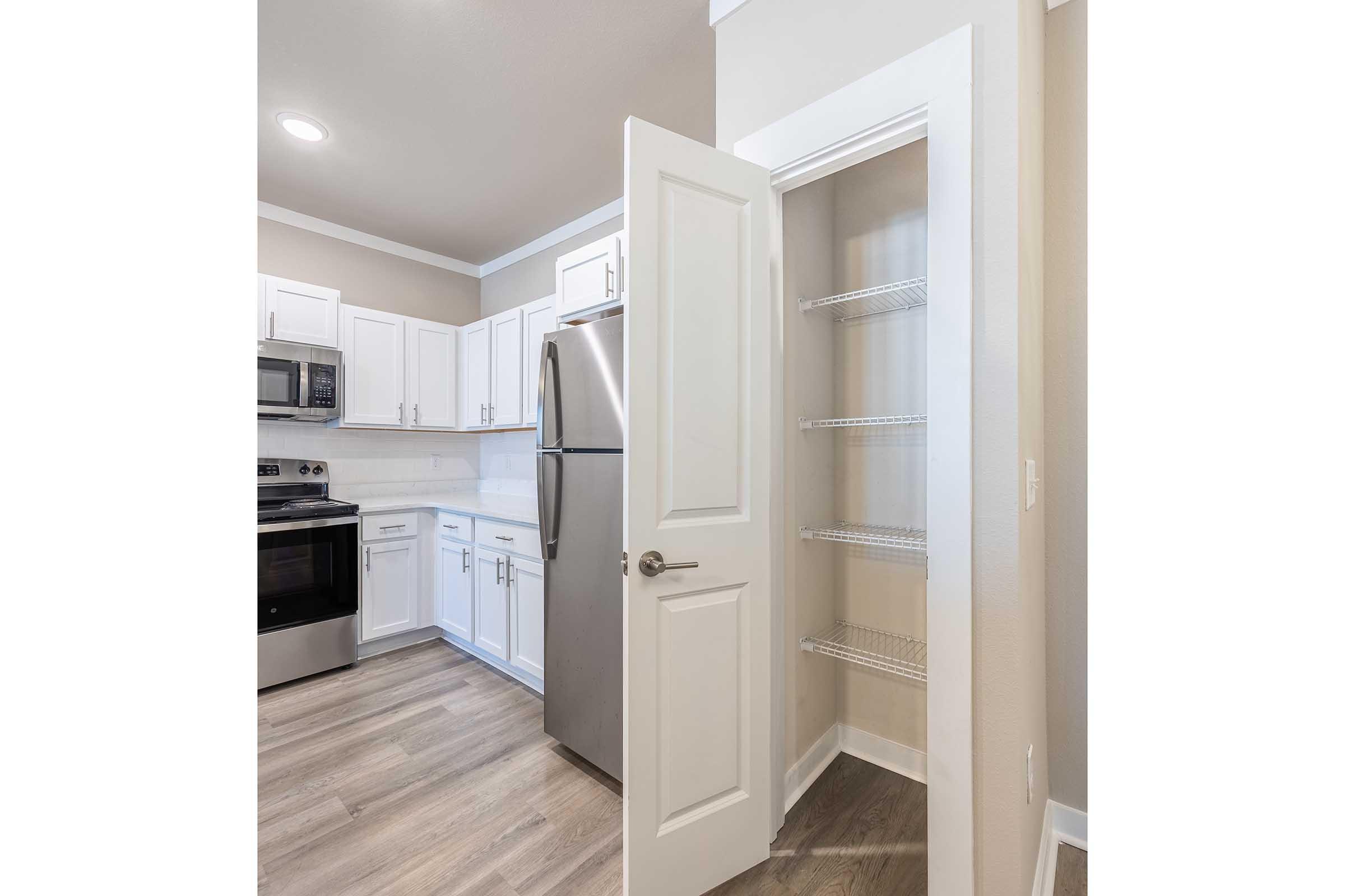 Modern kitchen featuring white cabinets, stainless steel appliances, and a pantry. The room has gray wood-like flooring and a neutral wall color. A door opens to the pantry with wire shelves, providing convenient storage space. Bright lighting enhances the clean and inviting atmosphere of the kitchen.