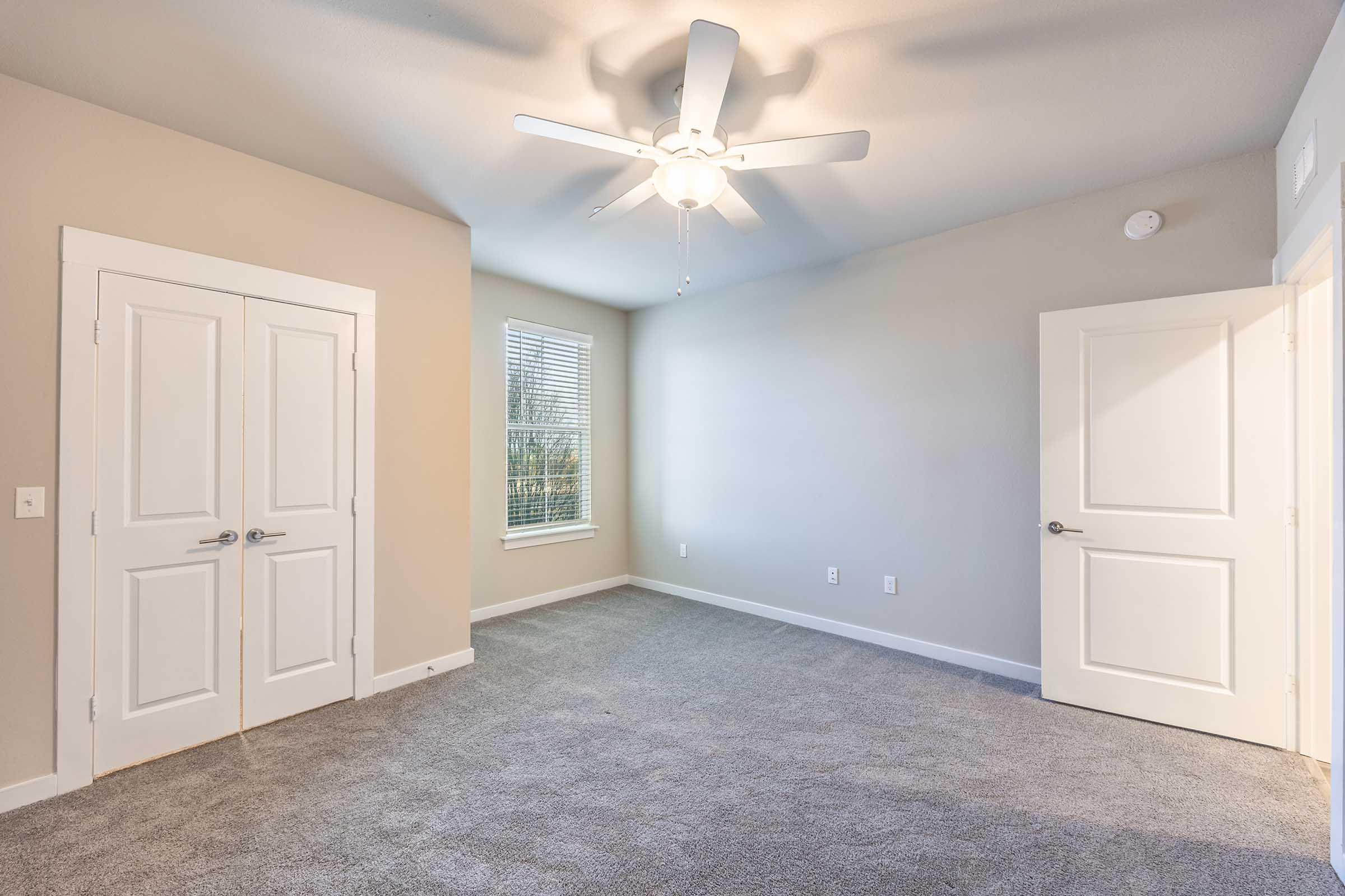 A spacious, unfurnished bedroom featuring light gray carpet, a ceiling fan, and two white doors. A window with blinds allows natural light, illuminating the neutral-colored walls. The room is empty, providing a versatile space for personalization.