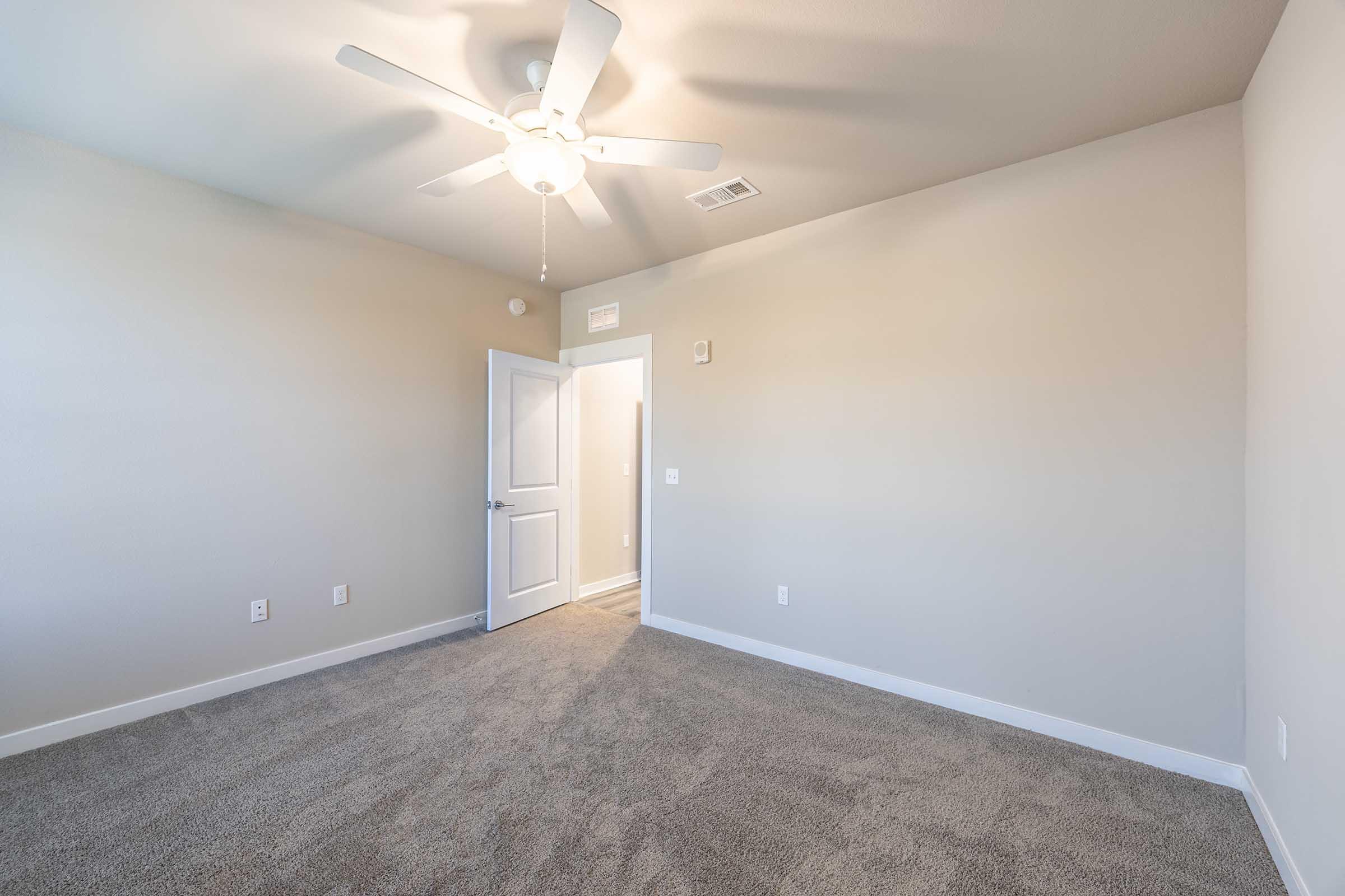 Empty room with light beige walls and carpeted flooring. A ceiling fan is mounted in the center of the room. A white door is slightly ajar, leading to a well-lit hallway. Natural light filters in, creating a spacious and airy atmosphere. Minimalistic design with no furniture present.