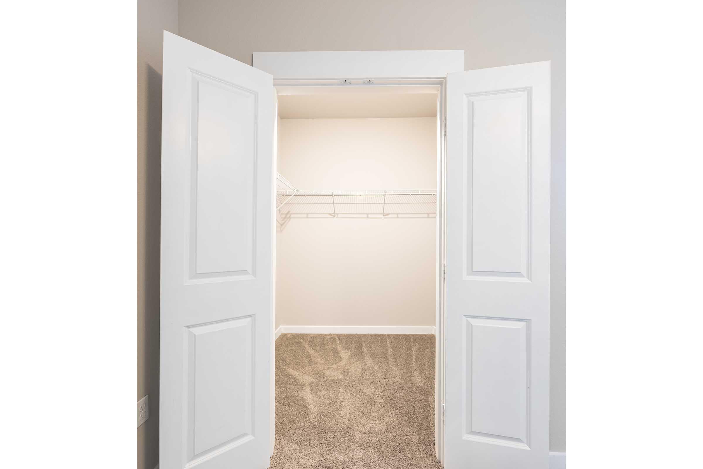 An open closet with white double doors, featuring a bare, light-colored interior and a wire shelving unit along the back wall. The floor is covered in soft, beige carpet, creating a clean and empty space.