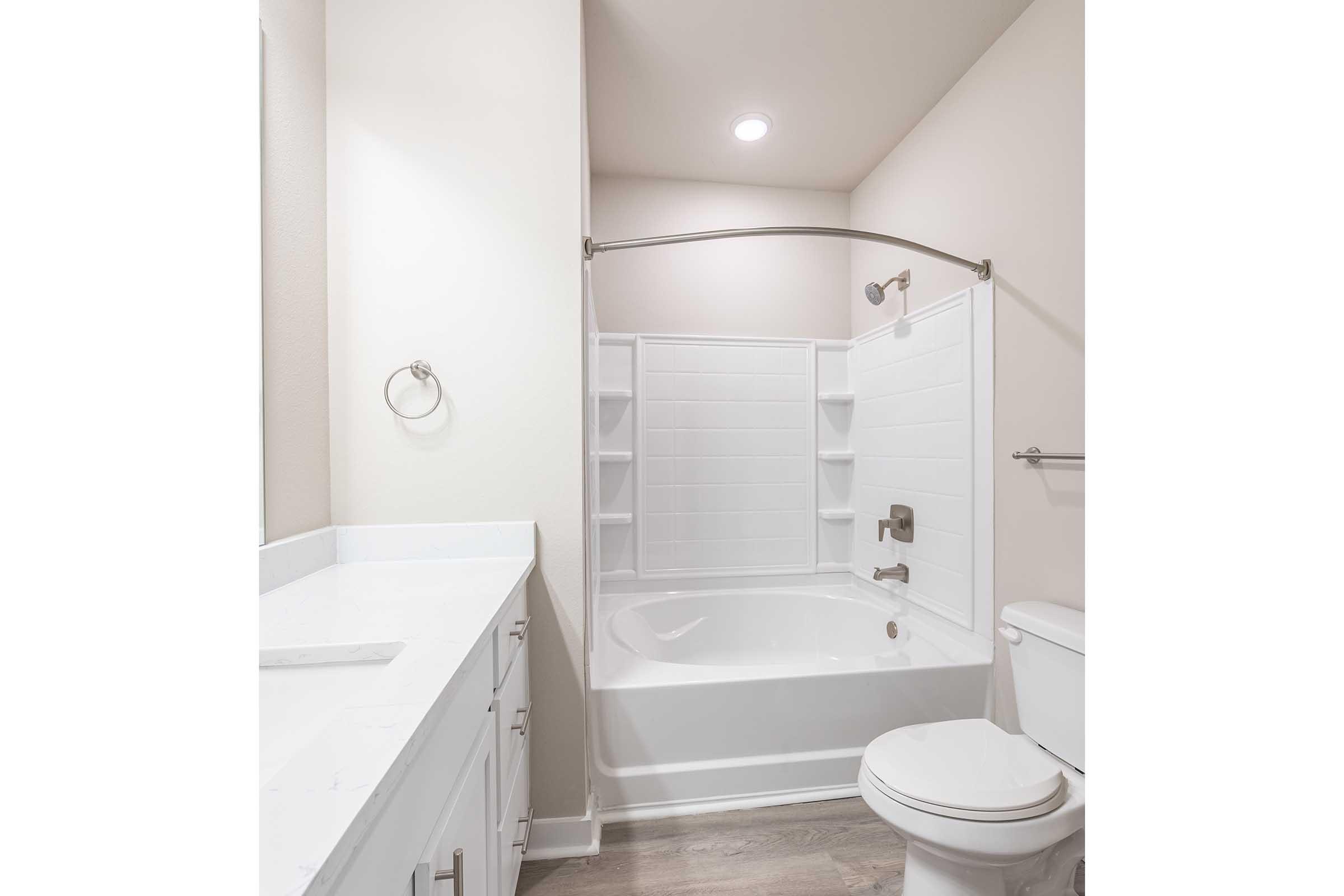 A clean and modern bathroom featuring a white bathtub with a curved shower rod and glass door, white tiled walls, beige paint, and a matching vanity sink with a mirror. The space includes a toilet and shelves for storage, with soft lighting illuminating the room.
