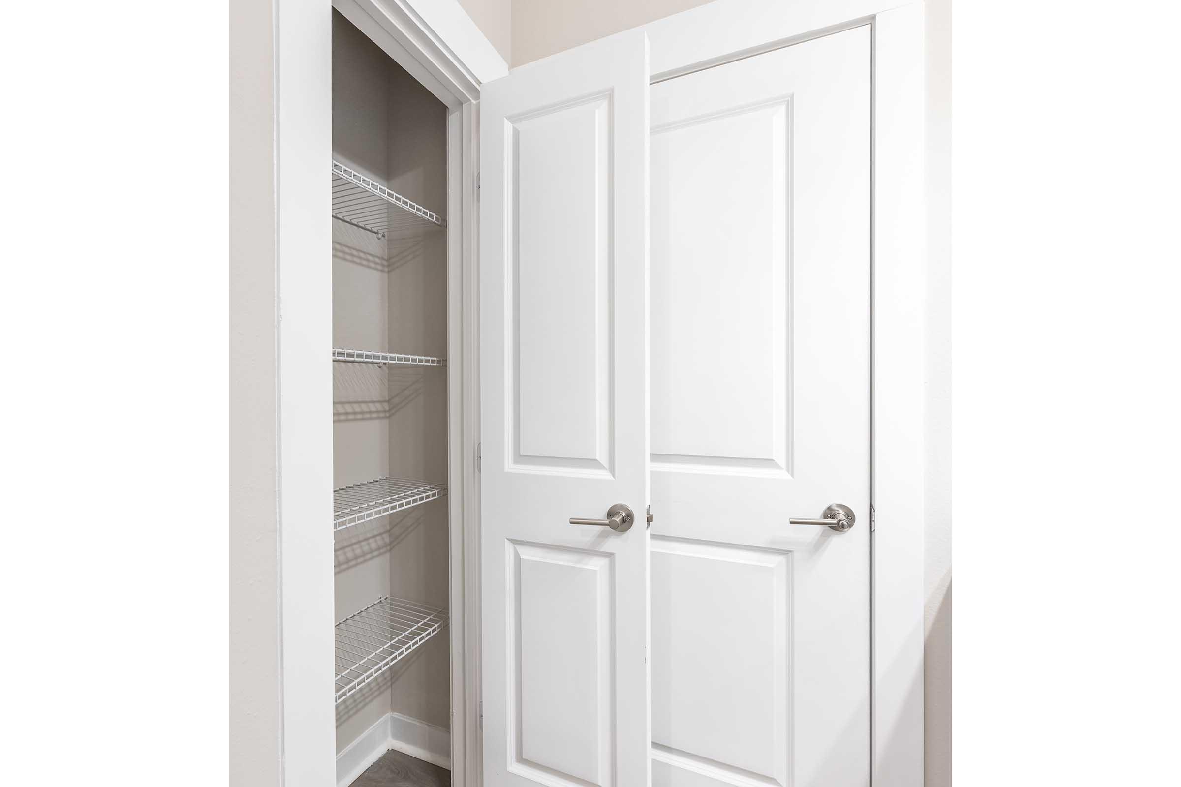 A view of a closet with double white doors slightly ajar, revealing empty wire shelves inside. The closet is well-lit with neutral-colored walls and a clean, uncluttered floor, creating a spacious appearance.