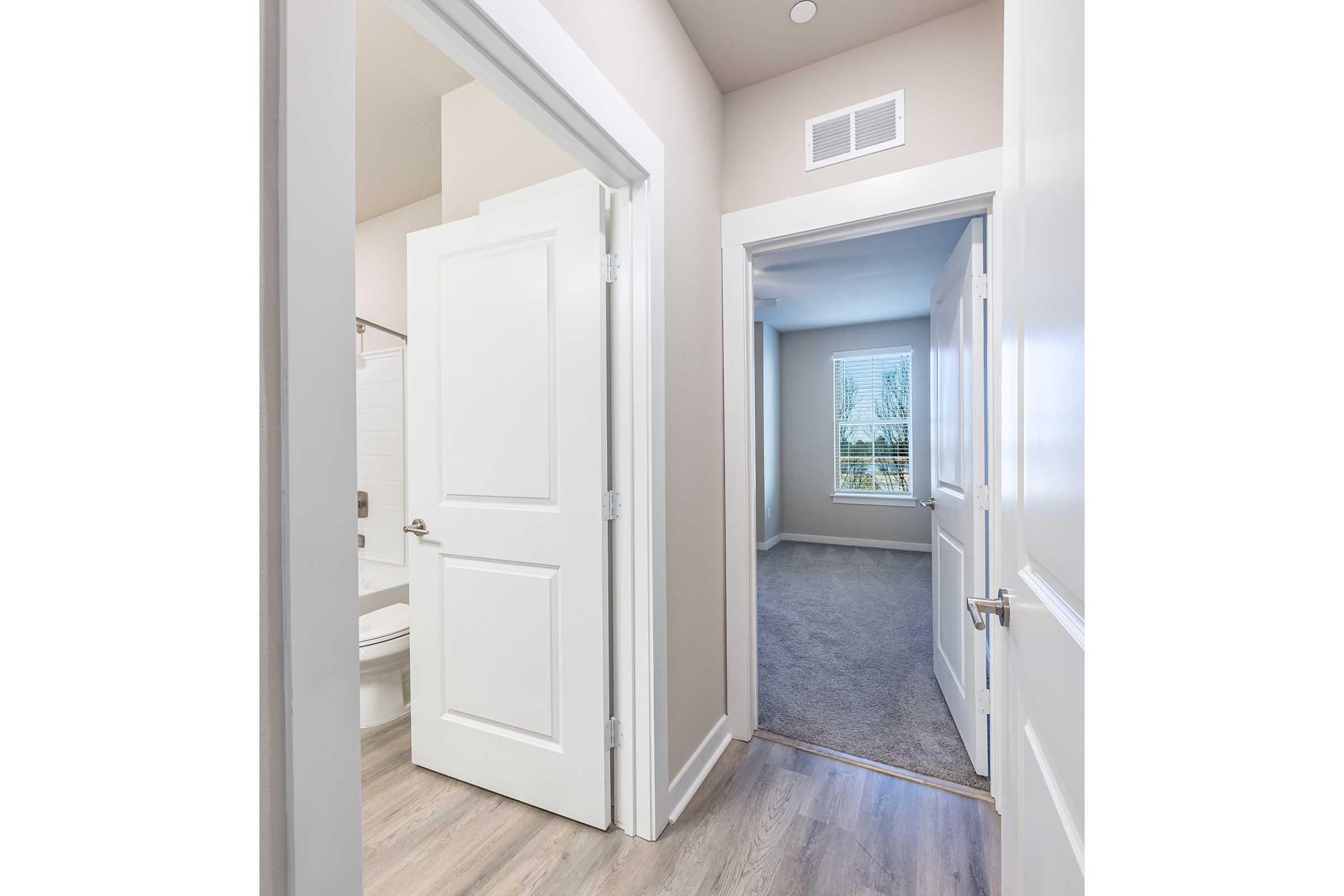 A hallway view showing two open doors. The door on the left leads to a bathroom with white tiles and a tub. The door on the right opens into a carpeted room with a window allowing natural light. The walls are painted in soft, neutral colors, enhancing the inviting atmosphere.