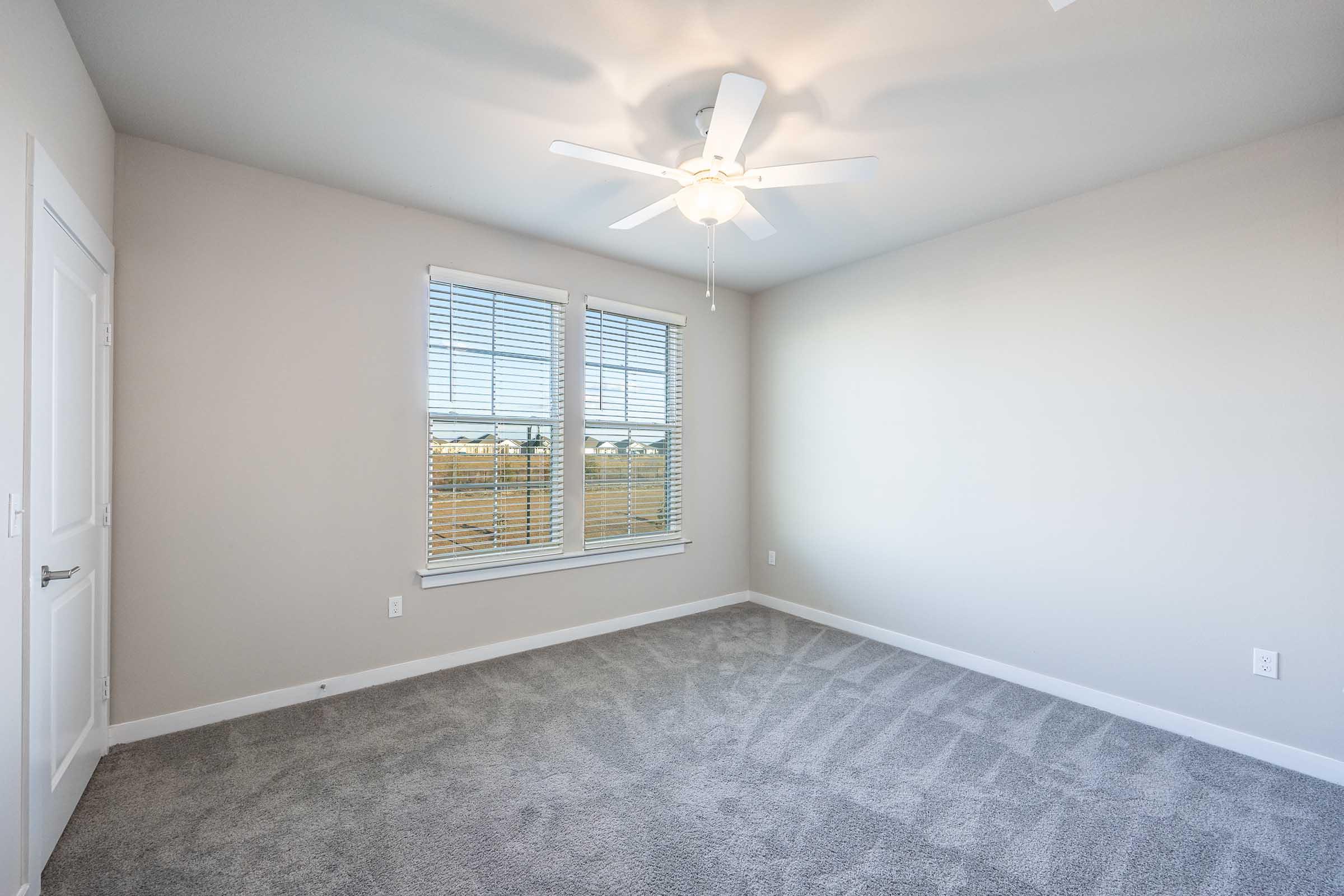 A vacant room with light gray carpet, featuring a ceiling fan and two windows with white blinds, allowing natural light to enter. The walls are painted a soft neutral color, and there's a door on one side. The overall atmosphere is bright and spacious, ready for furnishings.