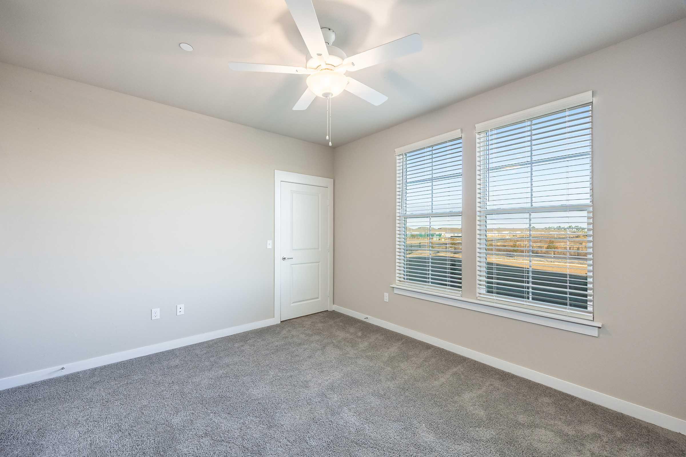 Empty room featuring beige walls, a ceiling fan with white blades, two windows with blinds letting in natural light, and a light gray carpeted floor. A white door is visible on the left side, and the room has a modern, clean aesthetic.