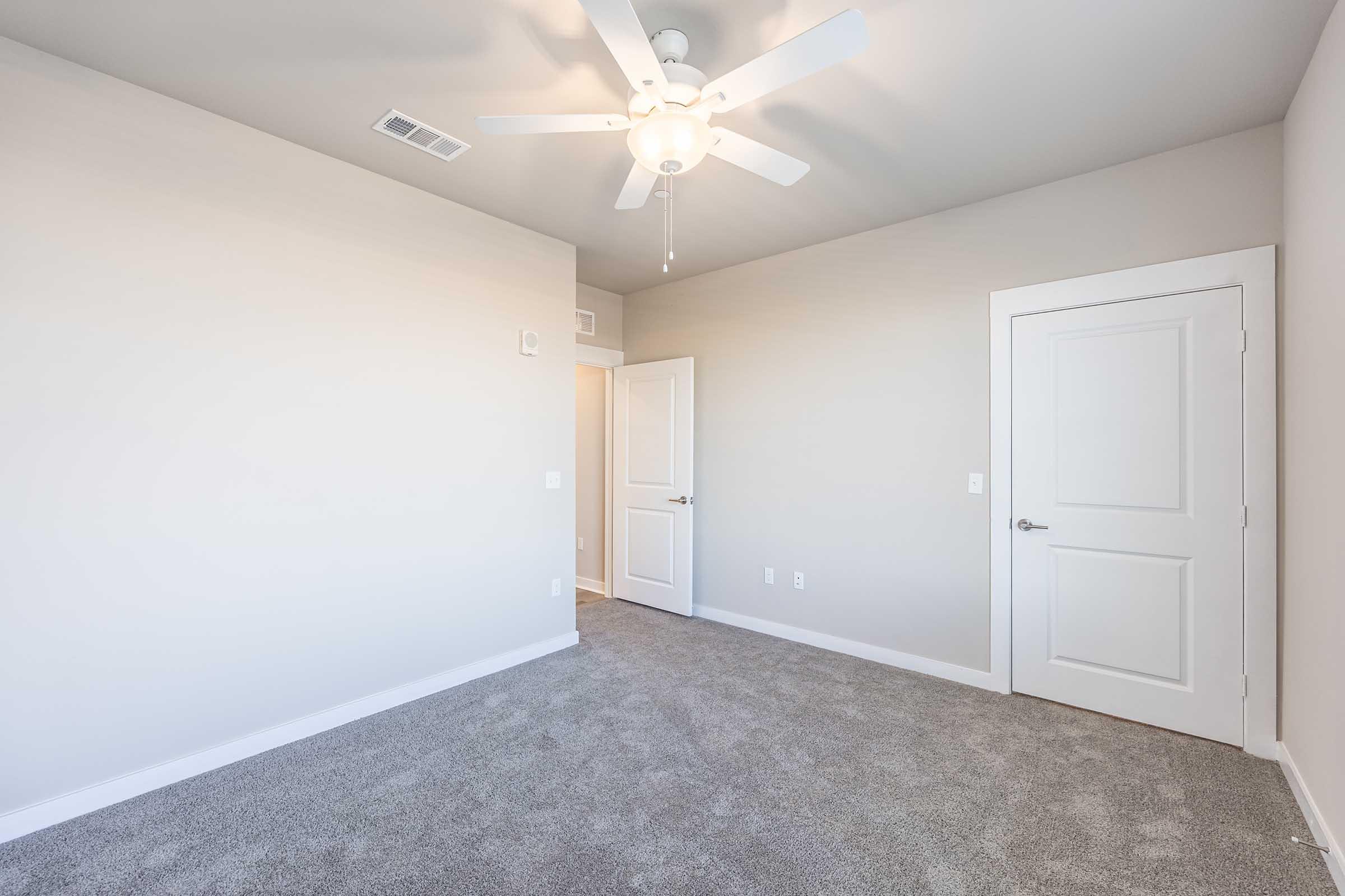 A well-lit, empty room with light gray walls and a ceiling fan. It features a plush carpeted floor and a closed white door on the right. The wall on the left shows no windows or decorations, emphasizing the room's spaciousness and simplicity.