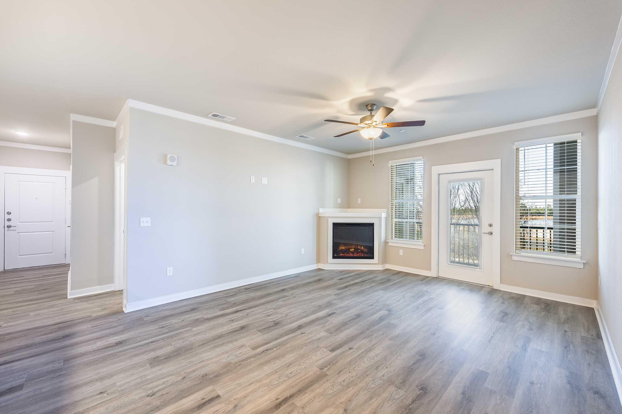 Spacious, well-lit living room with hardwood-style flooring, a ceiling fan, and a fireplace. Features large windows allowing natural light, a door leading outside, and neutral-colored walls. Ideal for creating a comfortable and inviting atmosphere.