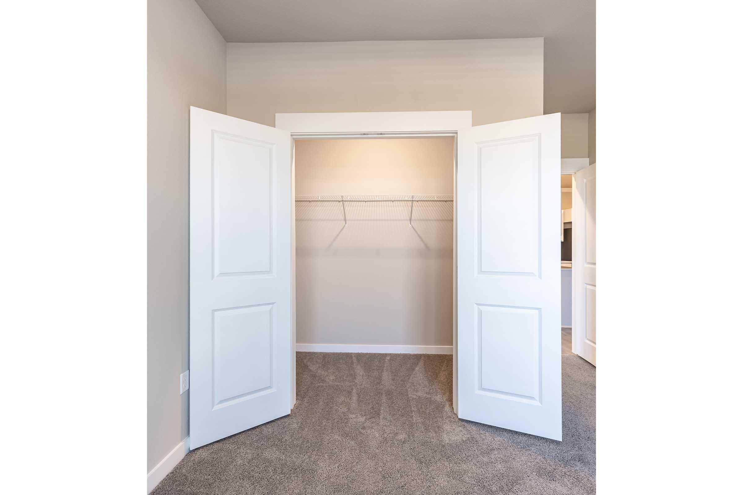 A spacious, empty closet with double white doors opened wide. The interior features a single wire shelving unit mounted on the back wall, and the floor is covered with plush, neutral-colored carpeting. The walls are a light, soft gray, creating a clean and airy feel.