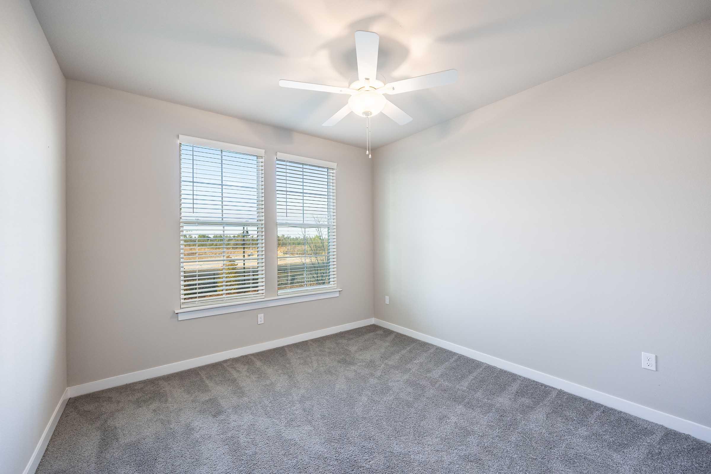 A spacious, unfurnished room with light gray carpet and beige walls. There's a ceiling fan and a large window featuring white blinds, letting in natural light and offering a view of outdoor greenery. The room is bright and airy, ideal for various uses such as a bedroom or office.