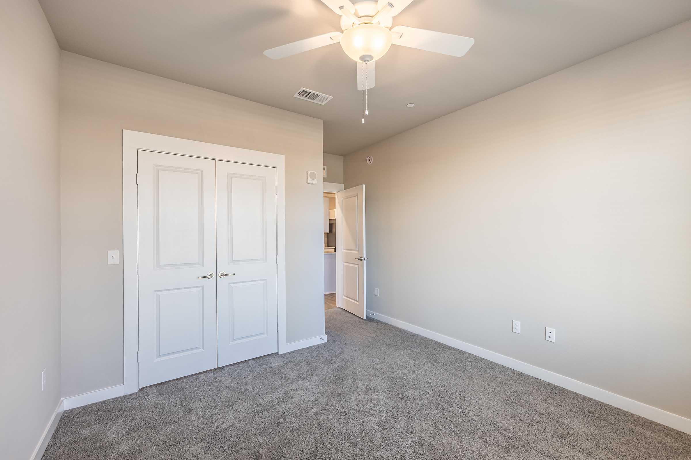 A light-filled, empty room featuring beige walls and carpet. There are double doors on one side leading to another room, while a ceiling fan is installed above. The space appears clean and modern, ideal for decorating or arranging furniture.