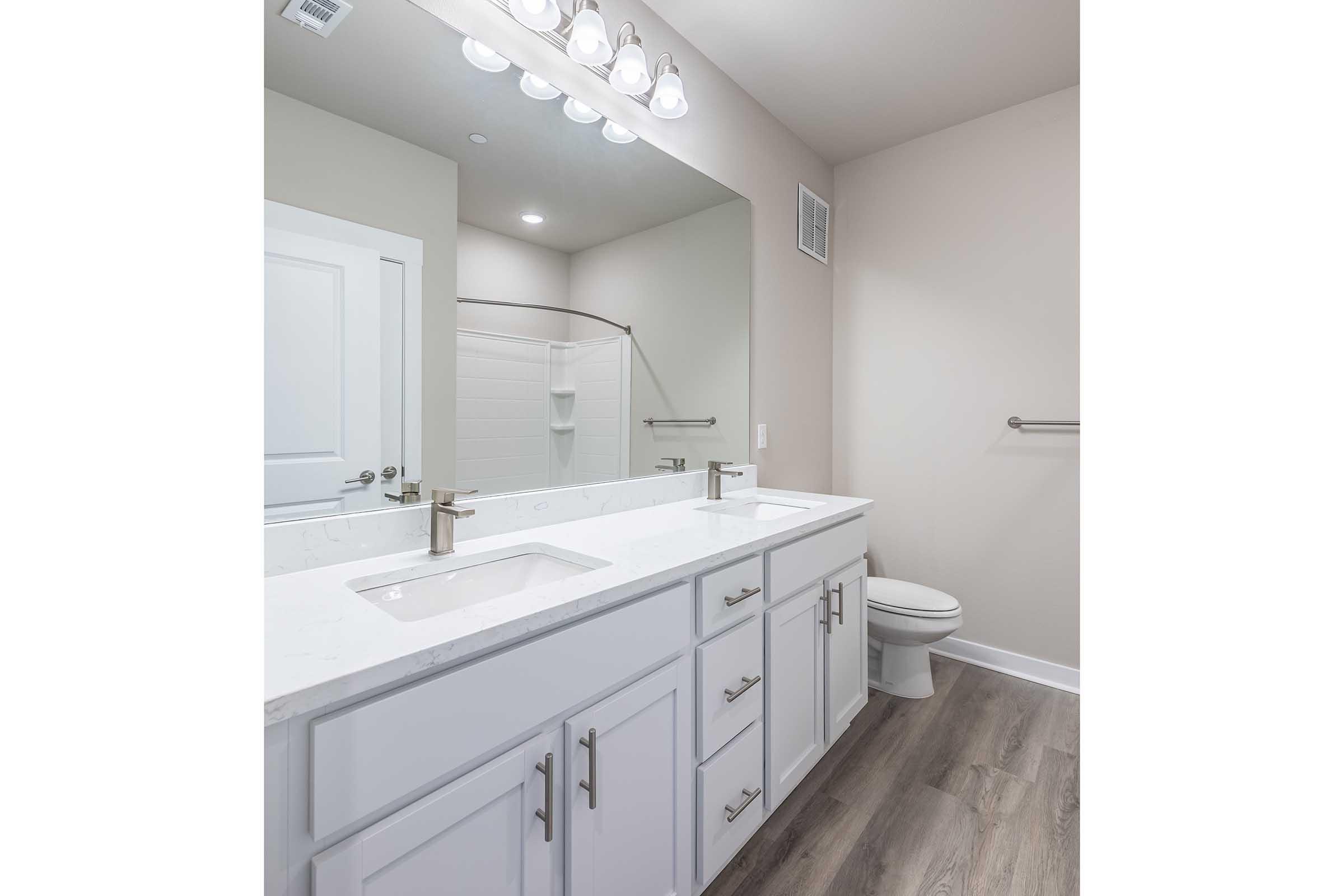 A modern bathroom featuring a double sink vanity with white cabinets, a large mirror, and stylish light fixtures. The space includes a shower area with a glass door, a toilet, and neutral-colored walls. The flooring is a light wood pattern, creating a clean and contemporary look.