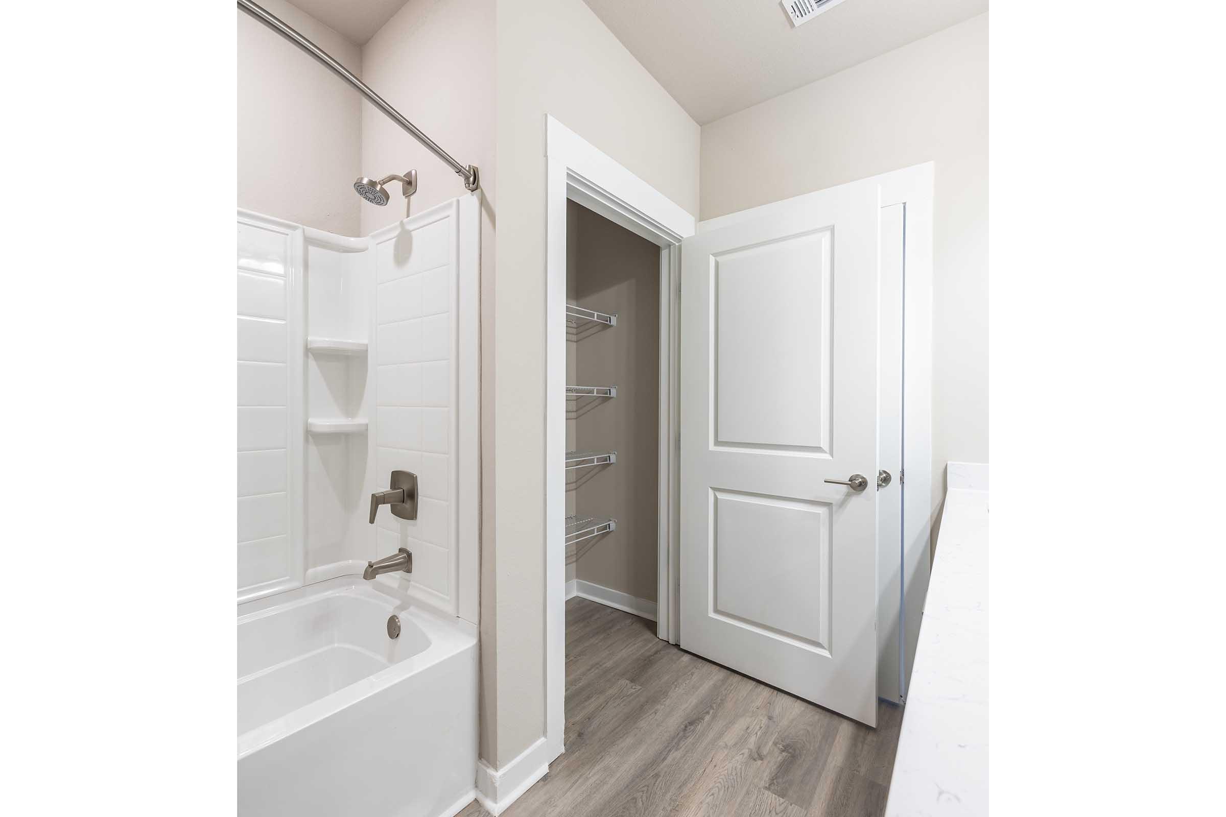 A clean and modern bathroom featuring a bathtub with a showerhead, beige walls, and light wooden flooring. On the right, there is a white door leading to a storage area with shelves. The lighting is bright, enhancing the spacious feel of the room.