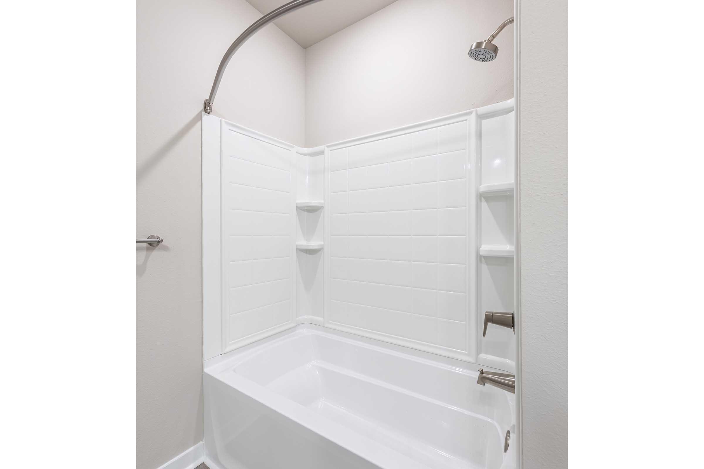 A clean, modern bathroom featuring a white bathtub with an attached shower. The shower has a curved curtain rod and a handheld showerhead. The tub area is enclosed with white tiled walls that include built-in shelves for toiletries. The overall design is minimalistic and bright.